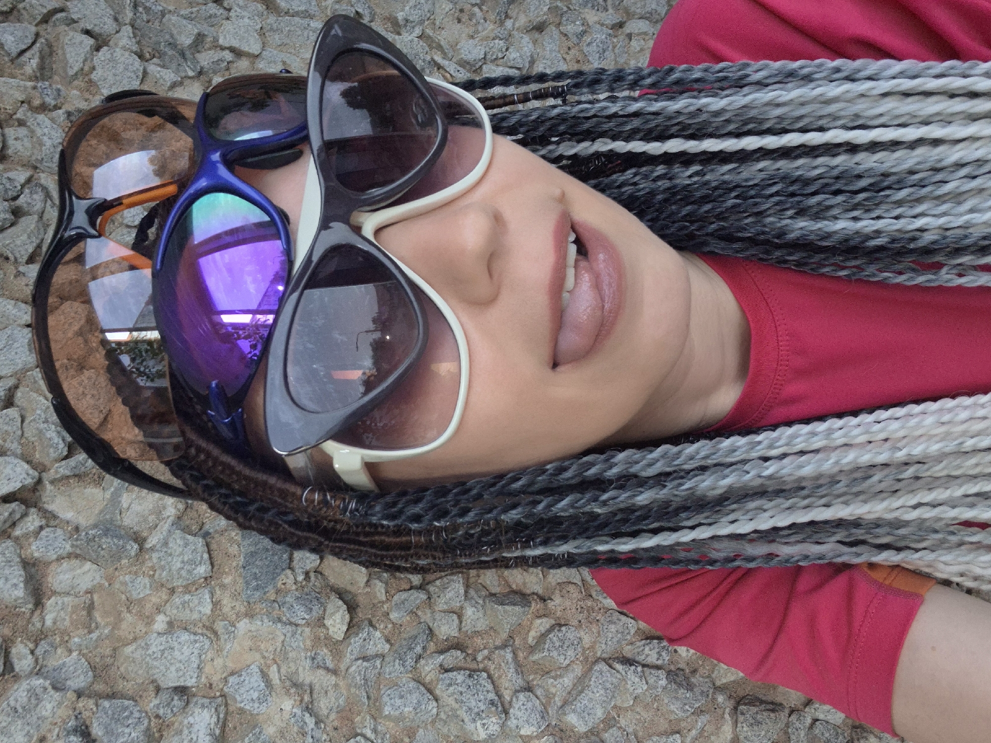 This media file shows a close-up, overhead view of a person lying on a stone surface. The person is wearing a bright pink shirt. They have long, braided hair with strands of black, grey, and white. The person is also wearing multiple pairs of sunglasses stacked on top of each other. One pair is blue with purple reflective lenses, another is black with white frames and dark lenses, and a third pair has brown lenses and black frames. The person's face is tilted upwards, with their mouth slightly open, revealing their tongue. The lighting suggests it is daytime, likely outdoors, given the stone ground and the reflections in the sunglasses. The overall mood appears casual and perhaps playful due to the way the sunglasses are stacked and the expression on the person's face. No visible text is present in the media file. The location context is Chaikovsky, Russia.