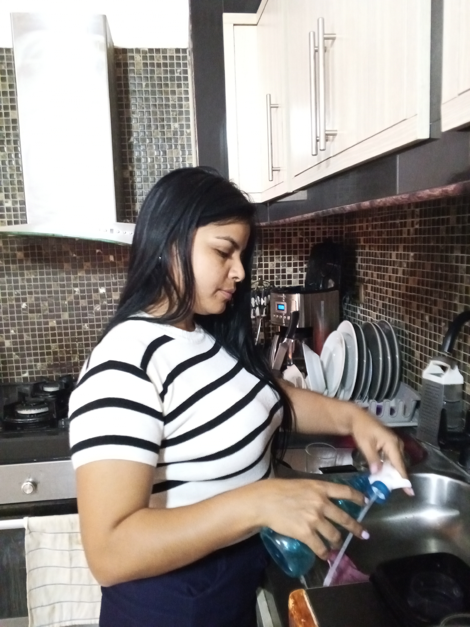 In a kitchen setting, a woman is positioned at a sink. She is wearing a white, horizontally striped shirt with black bands and dark pants. In her hands, she holds a blue spray bottle and appears to be dispensing its contents into the sink.
The kitchen environment includes cabinetry above the sink, covered in horizontal metal handles. To the left of the woman, a gas stove is visible, and behind her, the wall is covered in a mosaic-style backsplash. Further back, other kitchen appliances and dishes can be discerned.