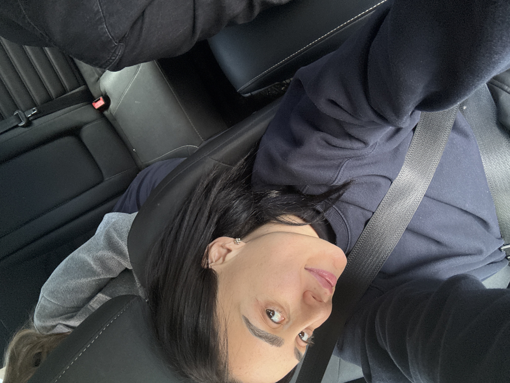 A woman with dark hair is seated in a car, wearing a navy blue sweatshirt and a seatbelt. Her head is tilted to the side, and she appears to be looking directly at the camera. The interior of the car is visible, including the black leather seats. The location cannot be determined from the image.