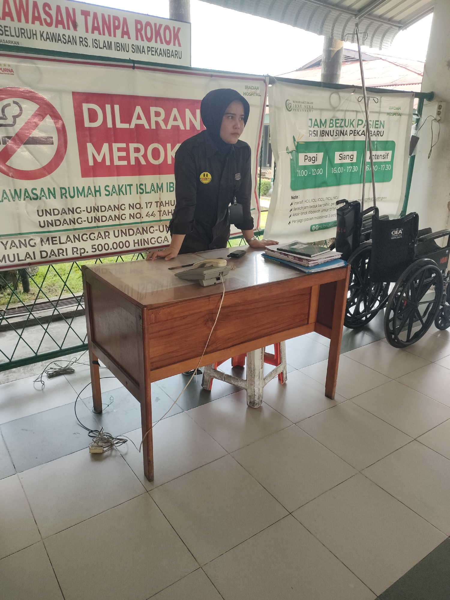 The image depicts an outdoor scene at the IBADAH HOSPITAL RSI IBNU SINA PEKANBARU in Pekanbaru, Indonesia. In the foreground, a woman wearing a black hijab and a black uniform with a yellow smiley face pin is standing behind a wooden desk. She appears to be an employee of the hospital, possibly an information desk attendant. On the desk are a landline telephone, a stack of books, and a mobile phone.
Behind the woman, a large banner with text in Indonesian is displayed. The banner prominently features a no-smoking symbol and the words "DILARANG MEROK" (Smoking is Prohibited). Another section of the banner lists visiting hours for patients, categorized into "Pagi" (Morning), "Siang" (Afternoon), and "Intensif" (Intensive). The text also mentions laws and regulations related to smoking, including fines.
To the right of the banner and woman, two wheelchairs are parked. The environment appears to be a walkway or waiting area, with tiled flooring and a simple covered roof overhead. The overall impression is that of a public area within a hospital setting, with clear signage intended to inform and regulate visitors.