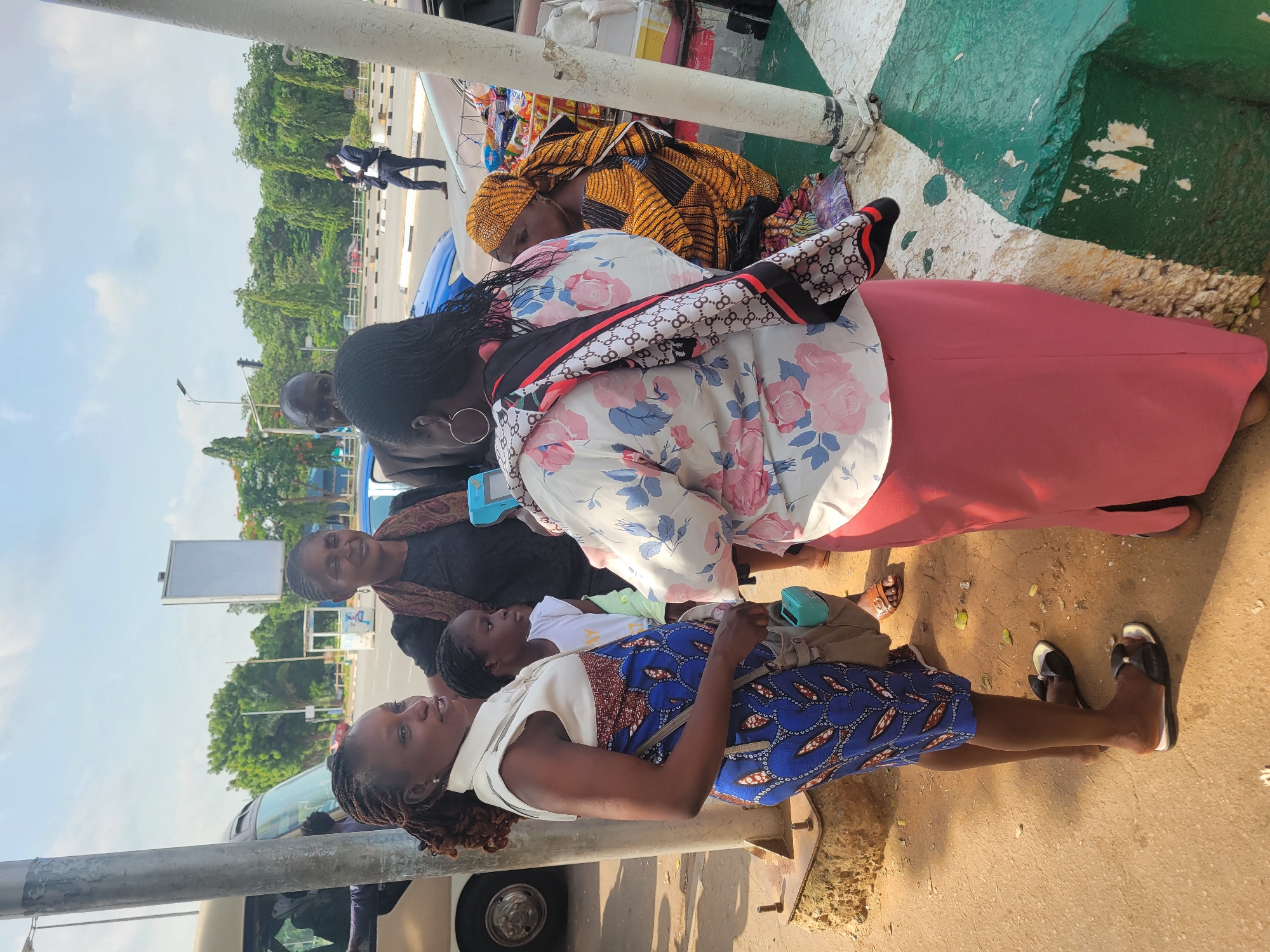 This vertical outdoor photograph captures a group of people, mainly women and a child, gathered in a public area under bright daylight, consistent with an urban setting in Abuja, Nigeria.
In the foreground, a woman with dark braided hair, wearing a white top and a blue patterned dress/skirt, stands facing right, holding a small light blue device. Next to her, a woman in a white floral-patterned top, a pink skirt, and a black and white scarf with a repeated "GG" logo looks downwards. Behind them, partially obscured, another woman is resting, wearing a vibrant yellow and black patterned headwrap and traditional attire. Various bags and personal items are visible around her. A third woman in a black top and patterned scarf smiles from behind the first woman, with a young child in a white t-shirt positioned between them.
The ground beneath them is a dusty, light-brown pavement. To the far right, a prominent structure painted in green and white with peeling paint suggests a connection to water, possibly a ferry terminal or dockside area. A sturdy white pillar or pole is visible in the immediate left foreground.
The background reveals a bustling urban landscape: a wide road with striped barriers, numerous lush green trees, and a clear blue sky with sparse clouds. In the distance, a man in a dark suit walks along the roadside, and several vehicles, including a blue car, are parked or passing by. An empty billboard frame stands tall in the mid-ground. The scene conveys a sense of waiting or transit in a busy city environment.