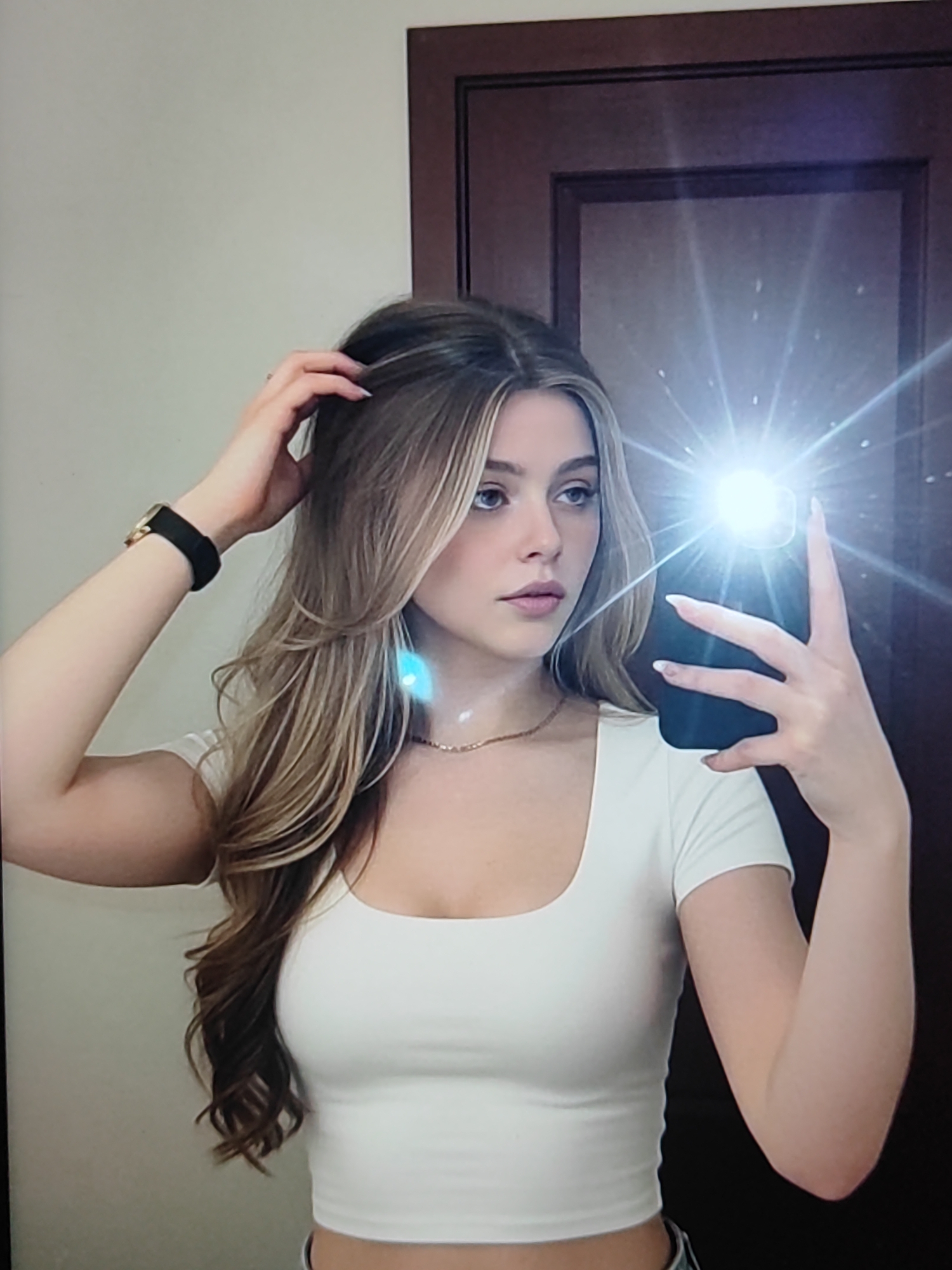 A young woman with long, layered blonde hair is taking a selfie in a room. She is wearing a white, cropped t-shirt and a delicate gold necklace. Her left hand is in her hair, and her right hand holds a black smartphone, with the flash illuminating the scene. The woman is looking towards the phone with a neutral expression.
Behind her, a dark wood door is visible, reflecting the bright light from the phone's flash. The wall to her left is a light, neutral color. The overall lighting suggests an indoor setting, possibly at any time of day given the artificial light. There is no visible text in the image.