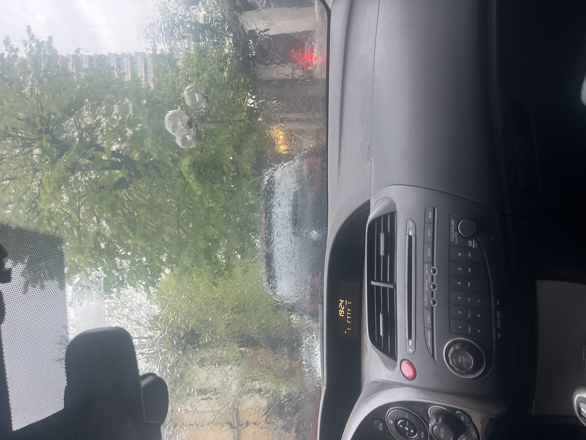 The image, captured from inside a vehicle, presents a view through a rain-streaked windshield, heavily obscured by water droplets. Inside the car, the dark grey dashboard is prominent on the right, featuring an audio system with an array of buttons, air conditioning vents, and a digital display. The display shows the time as "19:24" and includes partially visible characters "ΣΓΤΤΥ Σ". Controls for "TA/PTY", "CD/AUX", "AM/FM", "A.SEL", "SCAN", "RPT", "RDM", "TUNE/FOLD", "VOL", "SEEK/SKIP", "OP", "MODE", and "AUTO" are visible on the console, along with numerals 1 through 6. A portion of a dark headrest and the rearview mirror are visible on the left.
The scene outside the vehicle is blurred by the rain, revealing green trees with dense foliage, suggesting an urban or suburban environment in Varna, Bulgaria. A building can be vaguely discerned through the trees and rain. In the distance, red and yellow lights are visible, likely from another vehicle's taillights or turn signals, or a traffic light. A multi-globed street lamp is also faintly visible through the water on the glass. The weather is clearly rainy and overcast, corresponding with the evening time displayed. The car appears to be stationary, possibly parked or stopped in traffic, with no specific activity beyond observation of the weather.