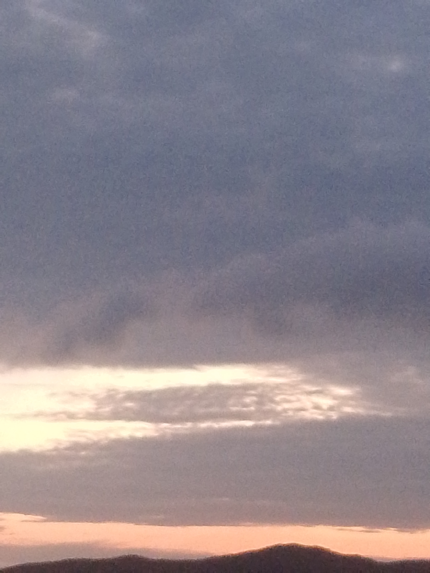 The image depicts a sky with a mix of gray and white clouds, suggesting an overcast or partly cloudy day. In the lower portion of the frame, there's a hint of land, possibly a mountain range, silhouetted against a pale horizon. The coloration suggests either sunrise or sunset. The absence of specific details like people, subjects, objects, or discernible actions prevents a description tied to Juan Griego, Venezuela.