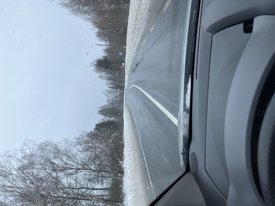 The image depicts a winter road scene from the interior perspective of a vehicle in Kopylovo, Russia. The foreground shows the top portion of a vehicle's dark gray dashboard and the upper edge of a steering wheel, with accumulated snow and ice visible at the base of the windshield. The windshield exhibits multiple small water droplets or smudges.
The primary subject is a two-lane asphalt road, wet with visible puddles and patches of ice, curving gently to the right in the distance. A continuous white line marks the center of the road, and faded dashed white lines are discernible in the right lane. Snow banks line both sides of the road, separating it from dense forestation. The forest consists primarily of bare deciduous trees and some scattered evergreen conifers, extending into the background.
The sky is overcast and gray, indicating a dull winter day. On the right side of the road, approximately mid-ground, a tall street light pole with an extended arm is visible. Below it and closer to the road, a blue rectangular sign with a white right-pointing arrow is affixed to a pole on the snow-covered shoulder. No people or other vehicles are visible in the scene.