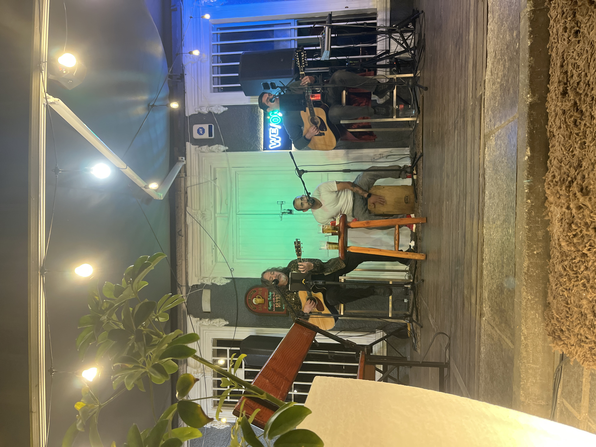 In Iquique, Chile, a band performs on a wooden outdoor stage illuminated by string lights and a green wash of light from a doorway. Three men are the subjects of the image. On the left, a man with long hair and a patterned jacket plays an acoustic guitar, seated on a stool. To his right, a man in a white t-shirt is seated and playing a cajon drum, with a microphone positioned in front of him. In the center and further back, another man in a grey shirt plays an acoustic guitar. He is seated on a stool, and a keyboard and other musical equipment are visible behind him. A neon sign spelling "WE ON" is visible in the background. Two speakers are positioned on either side of the stage. In the foreground, a large, roughly textured object, possibly a planter or a decorative element, is partially visible on the left. The scene appears to be a live music performance at a bar or restaurant.