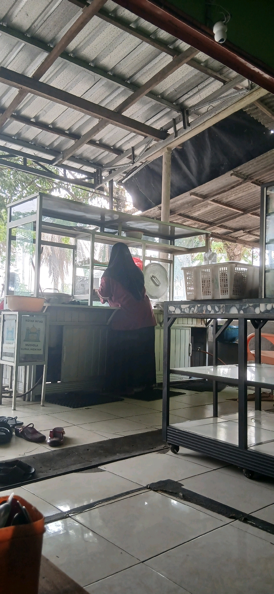 The image shows an outdoor food stall with a covered roof supported by metal beams and wooden planks. A person wearing a hijab and a maroon shirt is standing behind a counter, presumably preparing food. In front of the counter, there is a glass display case with a sign that reads "KOTAK INFAQ" and "MUSHOLA NURUL HIDAYAH," indicating a donation box for a prayer room. Several pairs of footwear are visible on the tiled floor, including sandals and flip-flops. The stall appears to be situated in a relatively open area, with trees and foliage visible in the background. The overall setting suggests a casual dining or service establishment in a tropical climate. The location is identified as Sumber Rejo, Indonesia.