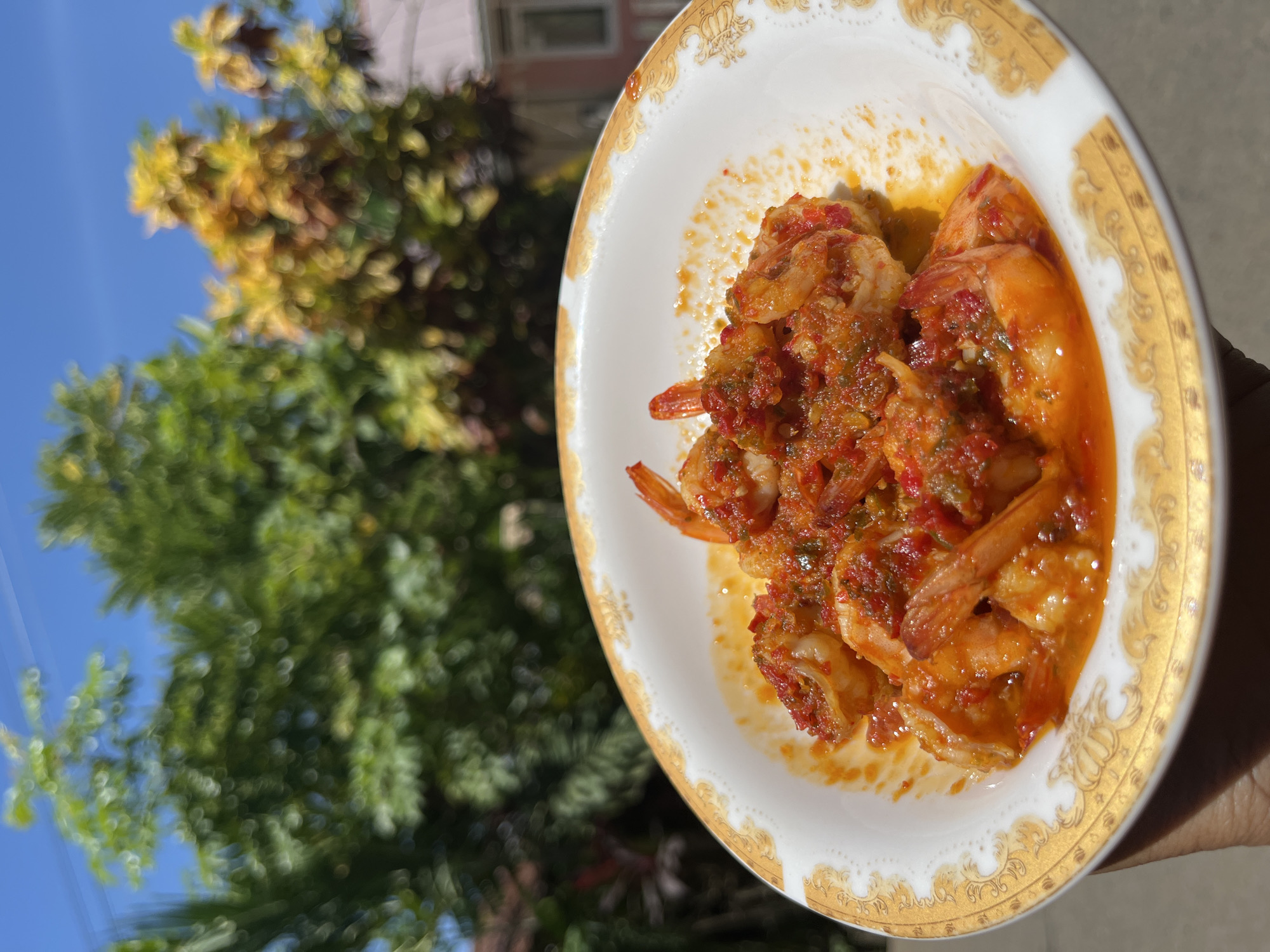 In a white ceramic bowl with a decorative gold rim, a serving of spicy shrimp is presented. The shrimp are coated in a vibrant red chili sauce, mixed with what appears to be garlic and other finely chopped ingredients. The bowl is held by a hand, partially visible at the bottom left of the frame. The background is blurred, showing lush green trees and a bright blue sky, suggesting an outdoor setting. A pink-hued building with a roof is also visible in the distant background. The image captures a meal in what is understood to be the city of Kruengmate, Indonesia.