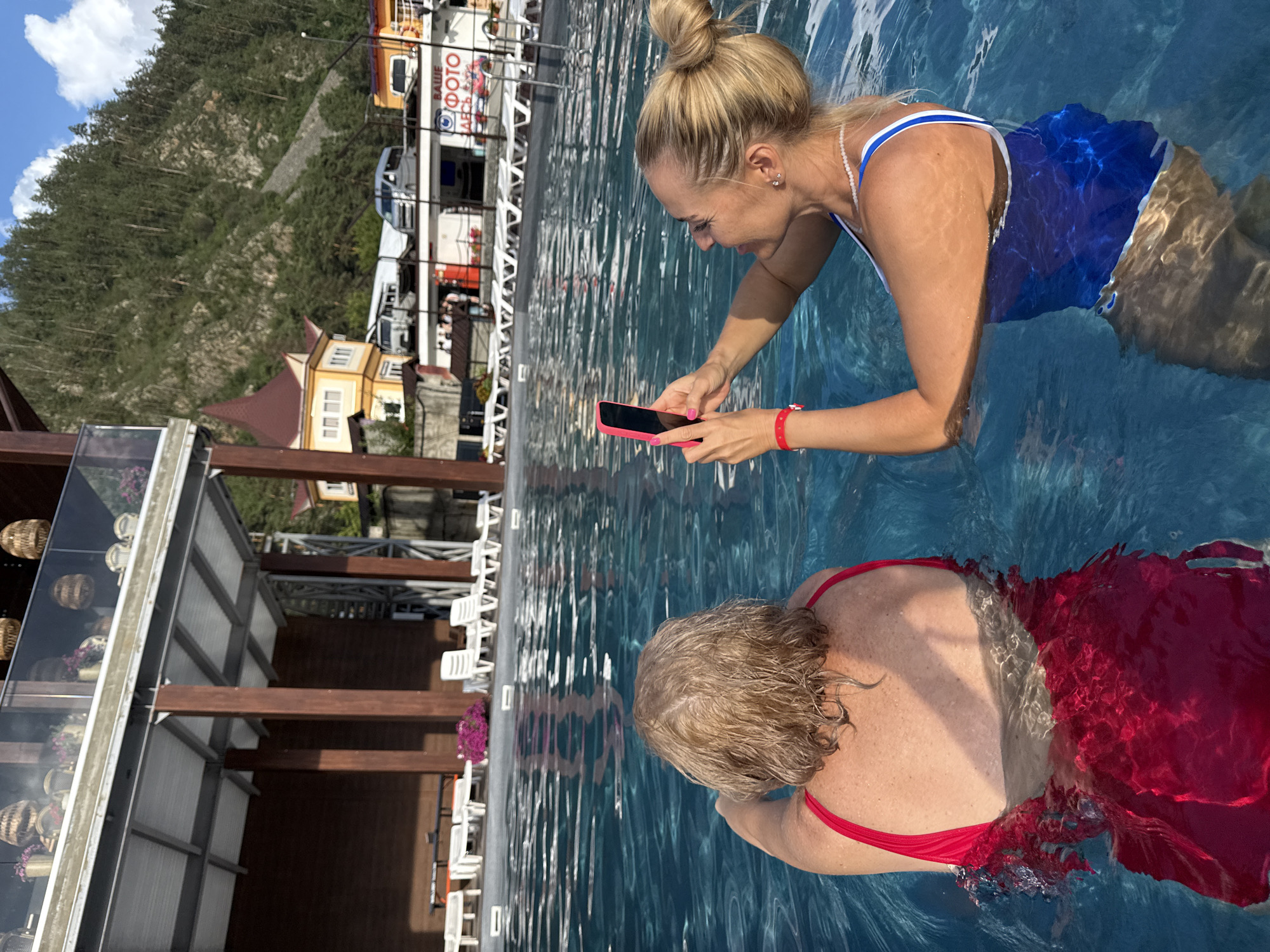 In Barangol, Russia, two women are in a swimming pool. The woman in the foreground on the right, facing left, has blonde hair pulled into a bun and is wearing a blue and white swimsuit. She is holding a pink smartphone and appears to be taking a photo or video. She is wearing a red bracelet on her left wrist and a pearl necklace. The woman in the foreground on the left is facing away from the camera, with blonde hair, and is wearing a red swimsuit. She is partially submerged in the water. The pool water is clear and blue. In the background, there is a building with wooden pillars and a balcony adorned with potted plants. Further back, there are lounge chairs, a yellow building with a red roof, and a forested mountainside under a blue sky with clouds. A sign with Russian text and a logo is visible in the background.