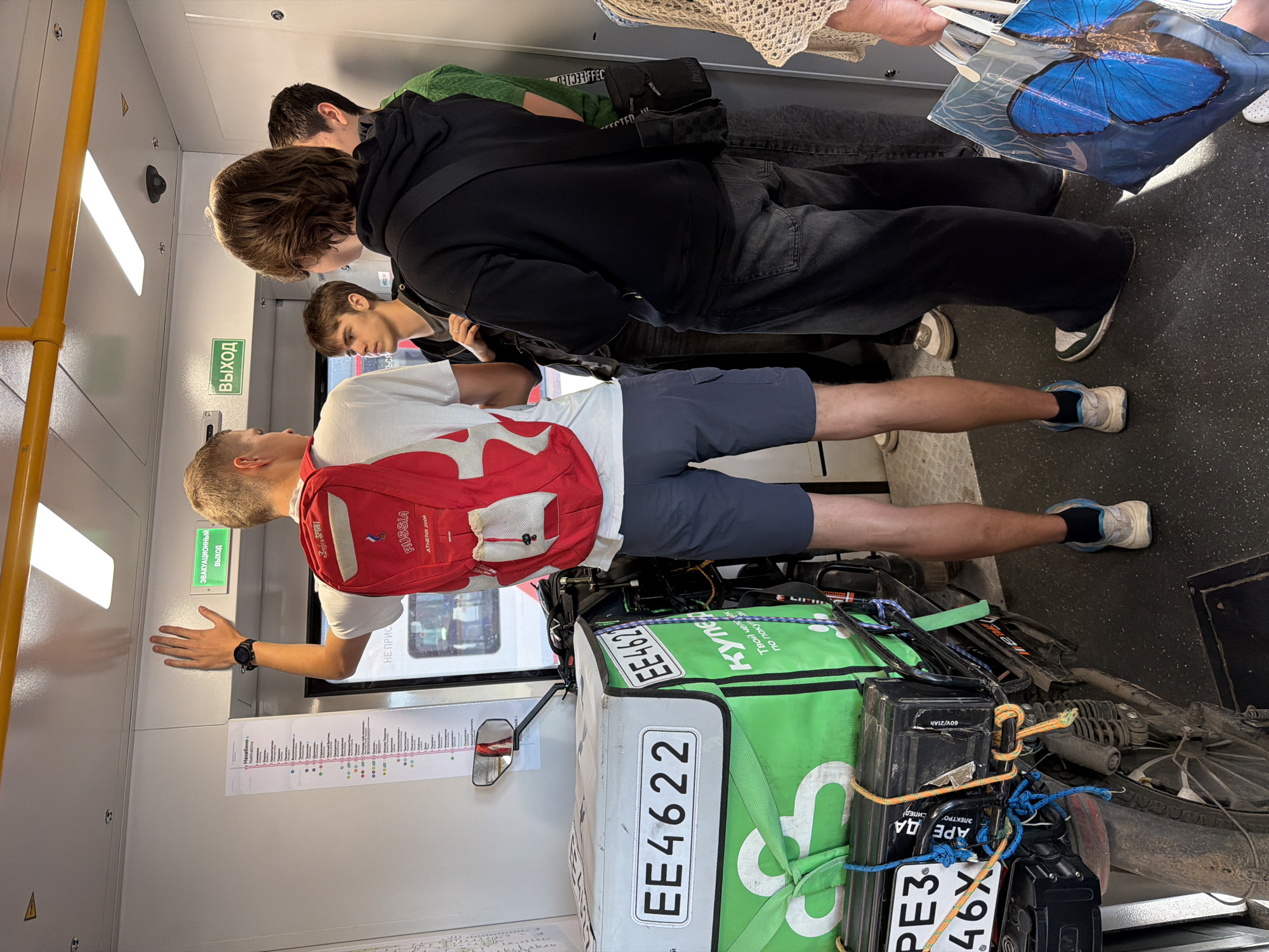 The image depicts a group of young people inside a train car, likely a metro or commuter train, in Moscow, Russia. The foreground is dominated by the rear of a motorcycle with a large, green delivery bag strapped to it. The bag has the text "EE4622" and a logo that appears to be "С". The motorcycle also has a license plate visible with the characters "РЕЗ" and "46X".
To the left of the motorcycle, a young man wearing a red backpack with the word "RUSSIA" and "ATHENS 2004" printed on it, is standing and reaching up with his right hand towards the ceiling. He is wearing a white t-shirt and blue shorts.
To his right, several other individuals are visible. A young man with brown hair is looking to the left, wearing a grey t-shirt. Behind him, another young man in a black hoodie and dark jeans is facing away from the camera. Further to the right, another person is partially visible in a green shirt.
On the wall of the train car, there is a route map and a green sign with the word "ВЫХОД" (Exit) in Russian. The train car interior appears functional and somewhat utilitarian, with yellow handrails along the ceiling and fluorescent lighting. The floor is dark grey. The overall scene suggests a moment of transit, possibly during a busy period.