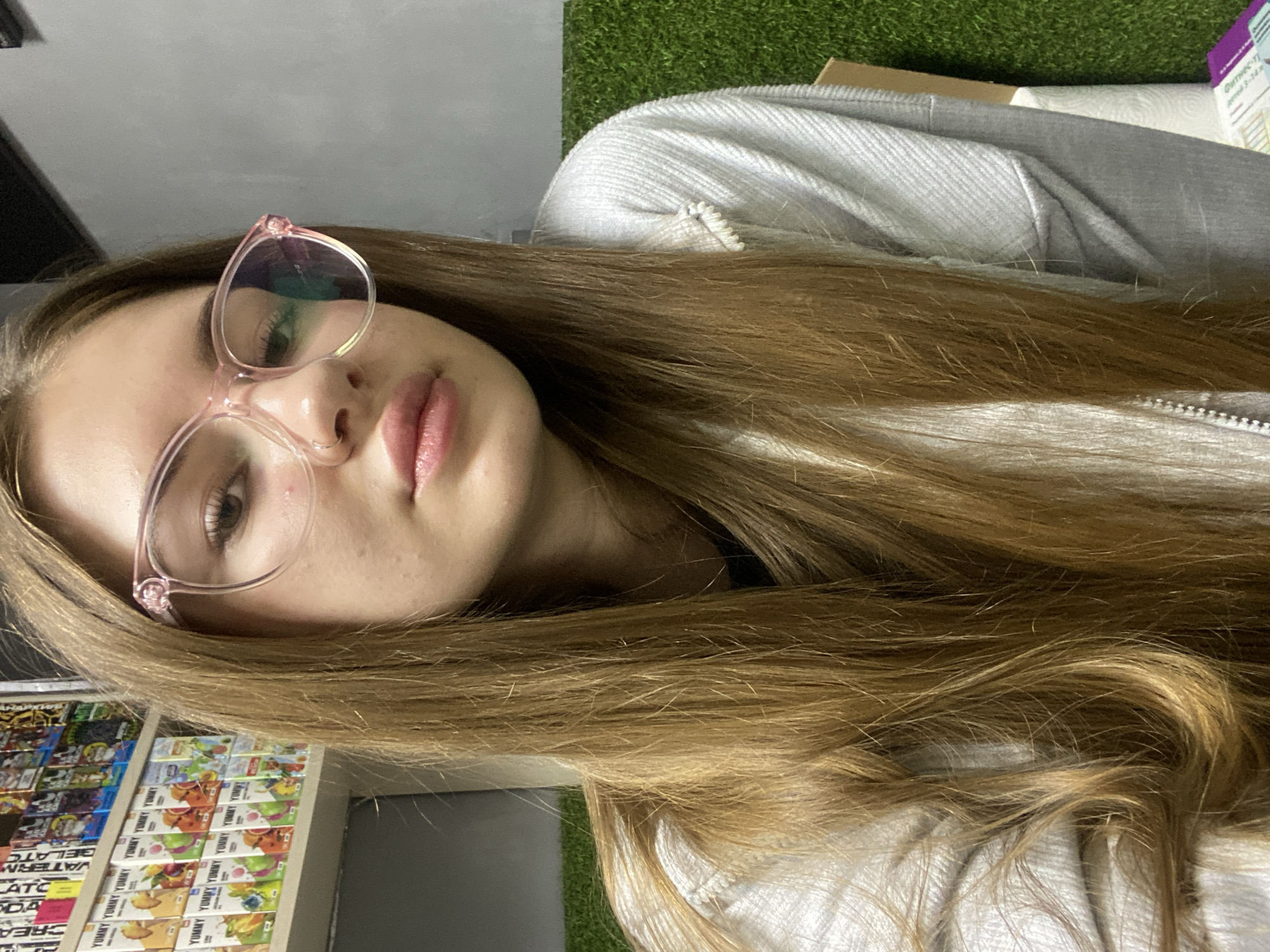 A young female with long, straight, light brown hair, wearing clear-framed pink eyeglasses and a light grey textured zippered jacket, is depicted from the chest up, looking directly at the camera. She has a subtle nose ring in her left nostril and full, light pink lips. The background features a commercial setting. To the subject's left, a multi-tiered shelving unit displays numerous small, rectangular packages branded "Yummy," depicting various fruit flavors such as watermelon, cherry, and blue razz, with both English and Cyrillic text visible. To the subject's right, the background consists of a plain grey wall in the upper section and a green artificial turf-like wall covering in the lower section. A brown cardboard box and a roll of white paper towels are partially visible in the bottom right corner against the turf. The image provides no specific visual information to confirm the location as Дьяково, Russia.