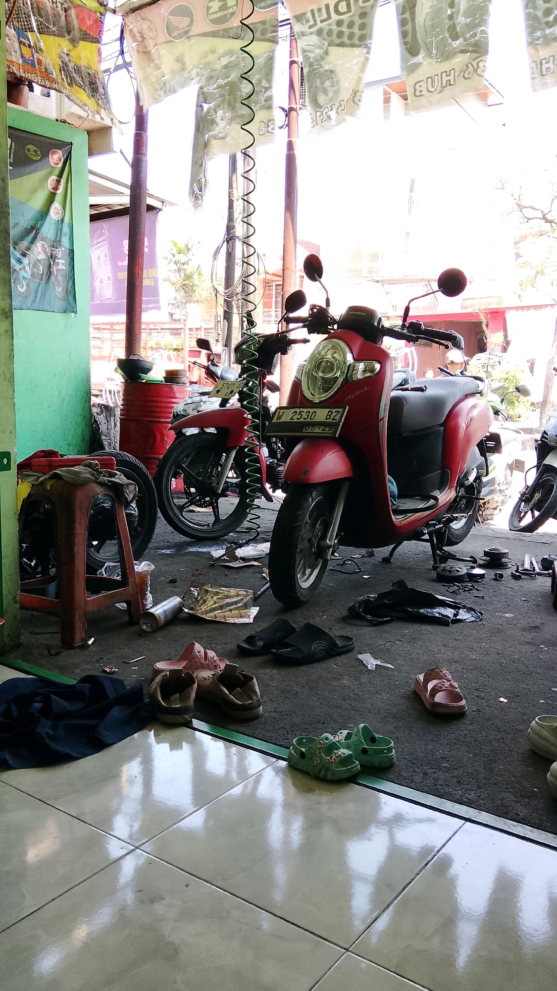 This is a photograph taken from a low angle, capturing the interior of what appears to be a motorcycle repair shop in Sukorame, Indonesia. The scene is cluttered with motorcycles, tools, and various debris. In the foreground, several pairs of sandals are scattered on the floor. A red stool is positioned to the left, with a piece of cloth draped over it. Two motorcycles are prominently displayed: a red scooter with a license plate reading "W 2530 BZ" and a red motorcycle behind it. The floor is a mix of concrete and tiled surfaces, with a prominent reflection on the tiles. In the background, banners with faded text and images hang from the ceiling. The overall impression is one of a busy, working environment.