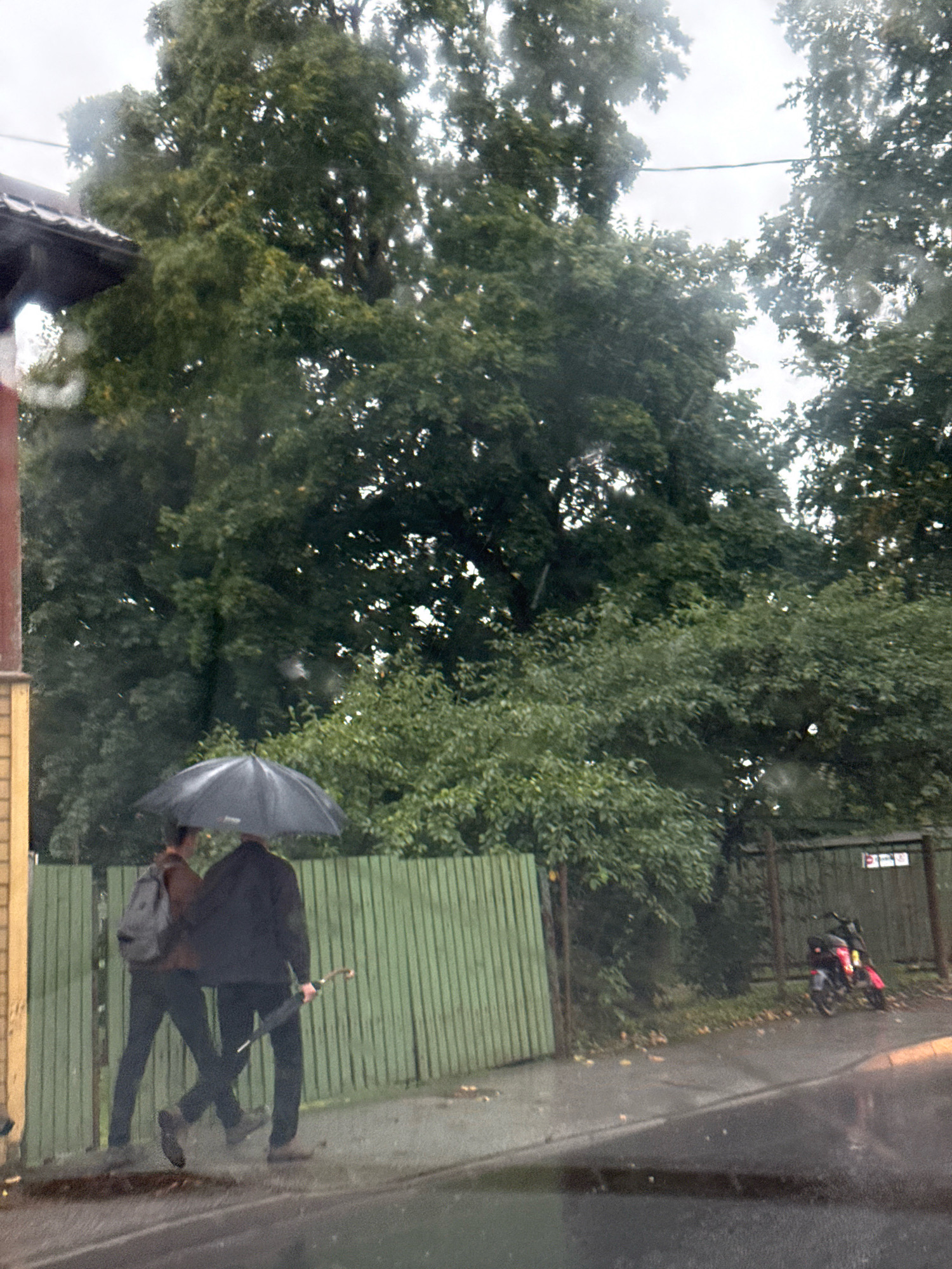 The image depicts an overcast day with rain, as indicated by water droplets on the camera lens and a wet pavement. Two individuals are walking on a sidewalk next to a green wooden fence, one holding a large black umbrella over both of them, and the other carrying a backpack and a folded umbrella. The background consists of lush green trees and foliage. To the right, a red and black motorcycle is parked near a metal fence with some signage. The scene appears to be a residential or suburban street in Tallinn, Estonia, judging by the architecture and vegetation.