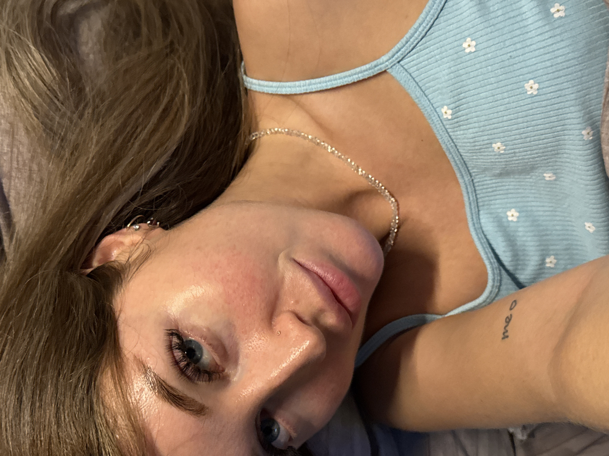 A young Caucasian female is captured in a close-up, overhead shot, lying down. Her brown hair is spread out around her head. She has striking blue eyes, accentuated by full eyelashes, and a subtle nose piercing. Her skin appears dewy and slightly flushed on her cheeks. She's wearing a light blue tank top with a delicate daisy pattern and a sparkly silver necklace. A small, handwritten tattoo reading "meo" is visible on her left forearm. The background is indistinct and appears to be indoors. No specific location in Chaikovsky, Russia can be determined from the image.