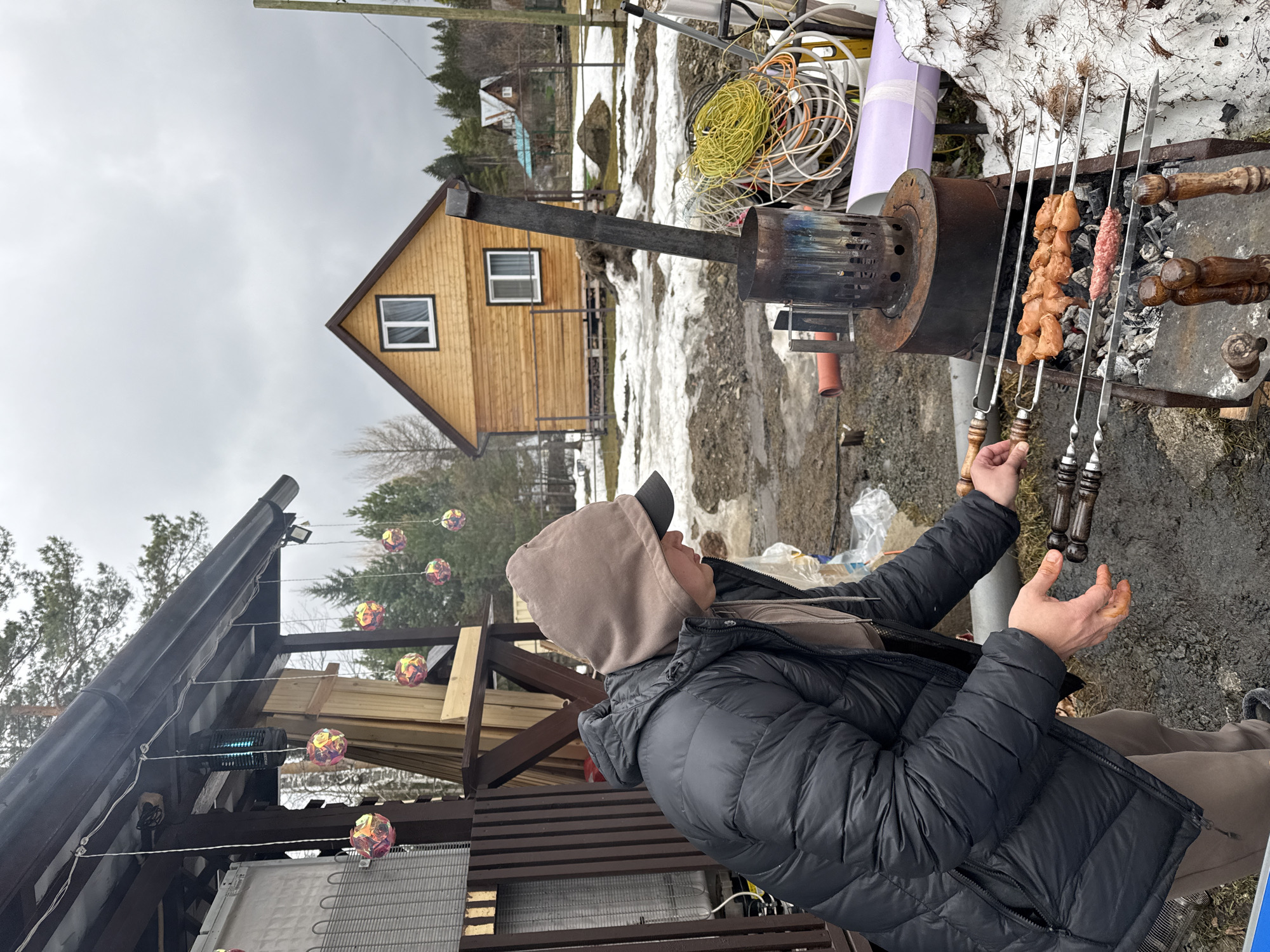 A man in a black puffer jacket and a beige hooded sweatshirt is standing outdoors, tending to skewers of meat grilling over a charcoal barbecue. The barbeque is a metal cylinder with a vertical chimney, placed on a stand next to remnants of snow. A wooden cabin is visible in the background, against a cloudy sky. Additional outdoor structures with lights and decor are partially visible to the left. The location is likely a residential or recreational area outside Yekaterinburg, Russia, evidenced by the vernacular architecture and snowy landscape, typical of the region.