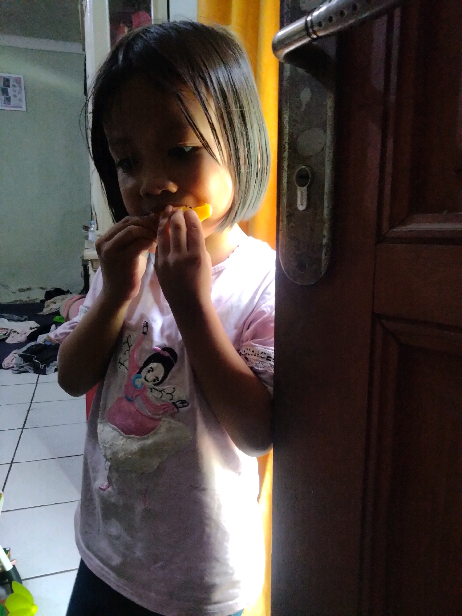A young child with dark hair is standing indoors, partially obscured by a doorway, and eating an orange piece of food. The child is wearing a light-colored t-shirt with a graphic of a character. The background shows a tiled floor, a light green wall with some clutter, and a yellow curtain. The doorway appears to be made of dark wood and has a metal door handle and lock mechanism. The lighting suggests the photo was taken during the day, with a strong light source coming from the right. The scene is likely a residential interior in Suka Sari, Indonesia.
