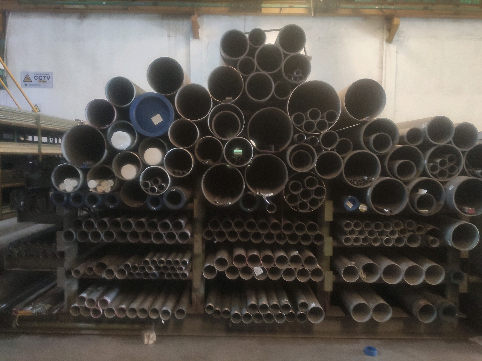 The image shows a warehouse filled with pipes. The pipes are of various sizes and are stacked on shelves. Some pipes are large and wide, while others are small and narrow. They are made of different materials, including metal and plastic. The pipes are organized in rows and columns on the shelves, with some pipes lying horizontally and others vertically. The warehouse has a concrete floor and walls, and there are some lights on the ceiling. In the background, there are some shelves with more pipes, as well as some other objects that are not clearly visible. There are no people or animals in the image. The overall impression is one of a well-organized storage facility for industrial materials.