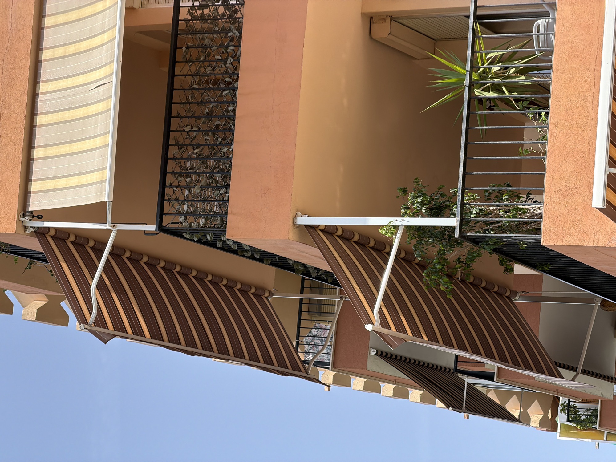 The image is an outdoor, low-angle shot of an apartment building with multiple balconies and retractable awnings in Palma de Mallorca, Spain. The building's facade is painted a warm, peachy-orange color. Several striped awnings are visible, some extended and others retracted. One extended awning features brown and tan stripes, while another higher up has lighter tan and white stripes. The balconies have black metal railings, and one balcony on the right contains a large yucca plant and a smaller tree-like plant. Another balcony further down on the left has a patterned cloth hanging on its railing. The sky is a clear, bright blue. The architectural style suggests a Mediterranean setting.