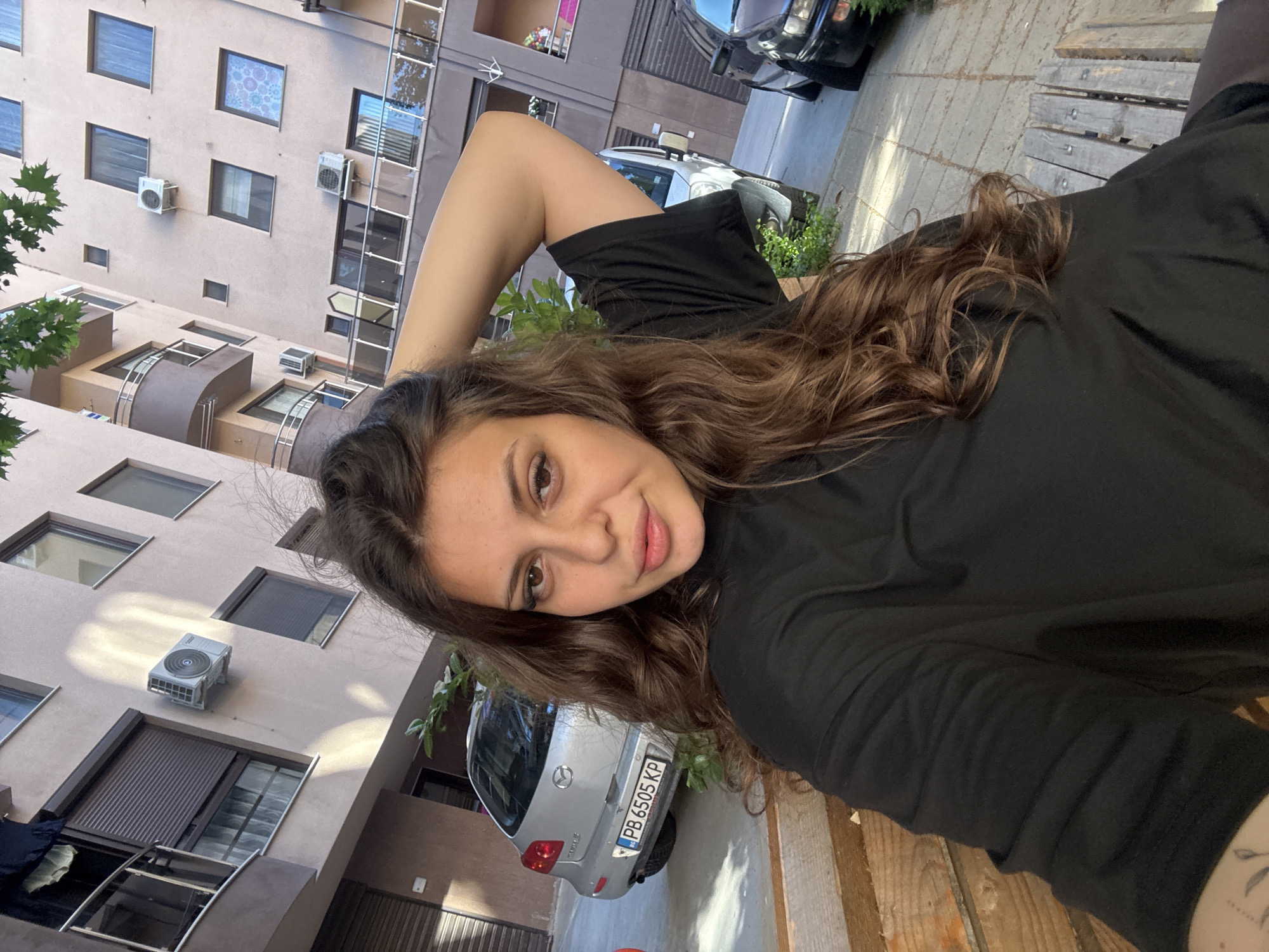 The image shows a young woman with long, wavy brown hair, wearing a black t-shirt, sitting on a wooden bench. She is looking at the camera with her left arm raised behind her head. In the background, there is a modern apartment building with balconies and windows, a silver Mazda car with a Bulgarian license plate (PB 6505 KP), and another car to the right. The scene appears to be in an urban setting, likely a residential area in Plovdiv, Bulgaria, given the Bulgarian license plate. The lighting suggests it is daytime.