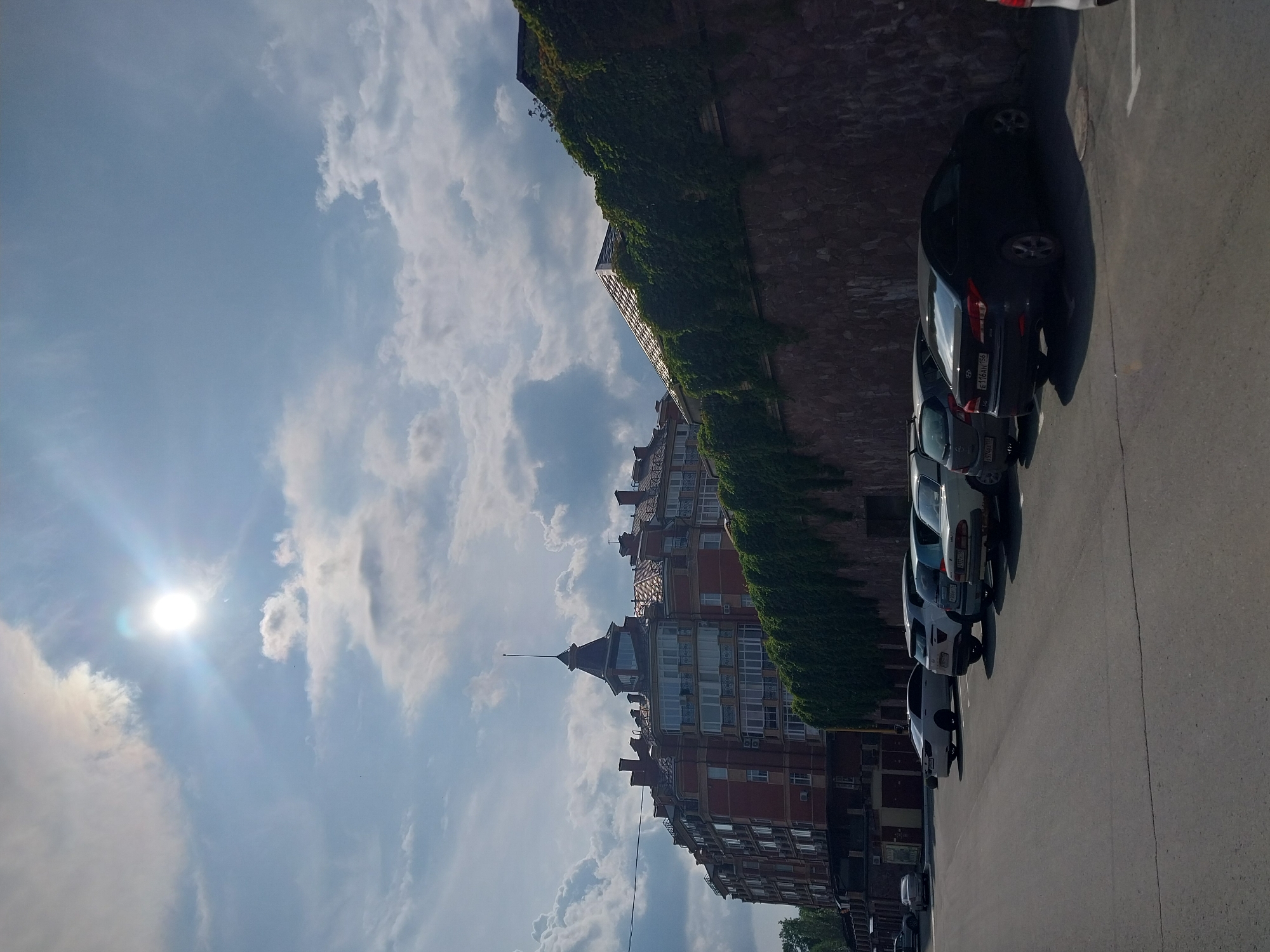 The image depicts an outdoor scene in Orenburg, Russia, likely during the daytime given the bright sun and blue sky with scattered clouds. The perspective is tilted, giving the impression of looking upwards.
The foreground features a row of parked cars on an asphalt surface. The cars are of various colors, including black, white, and silver. License plates are partially visible on some vehicles.
Behind the cars, a multi-story building with red brick walls and numerous windows is visible. The building has a distinctive architectural style with decorative elements and a prominent spire-like structure. Lush green ivy covers a section of the wall adjacent to the building.
The sky is a prominent feature, with a bright sun casting a lens flare and radiating light. Puffy white clouds are scattered across the blue expanse. The overall impression is one of a sunny day in a European-style urban environment. No people are clearly visible in the image. No discernible text is legible in the image.