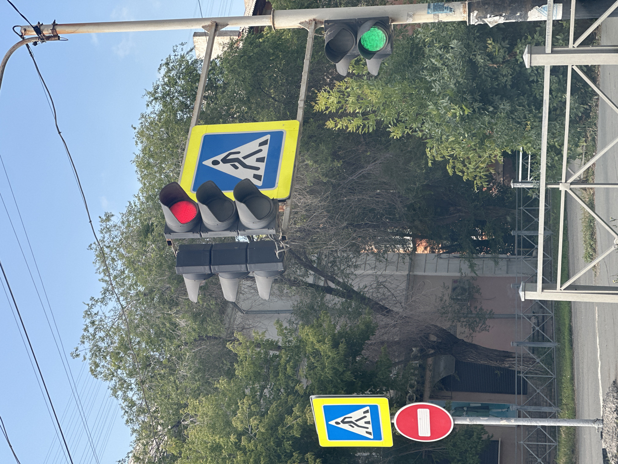 The image depicts a street scene in Orsk, Russia, featuring a traffic light with a red signal illuminated, indicating a stop. Below the traffic light, a blue square sign with a yellow border displays a white pedestrian symbol, signifying a pedestrian crossing. To the left of this setup, a red circular sign with a white horizontal bar indicates "no entry" or "do not enter." The traffic light on the right side of the image shows a green signal, suggesting that traffic can proceed in that direction. The scene is set against a backdrop of trees and buildings, with overhead power lines visible in the sky. The presence of these traffic control signs and lights suggests an intersection or a pedestrian crossing area within the city.