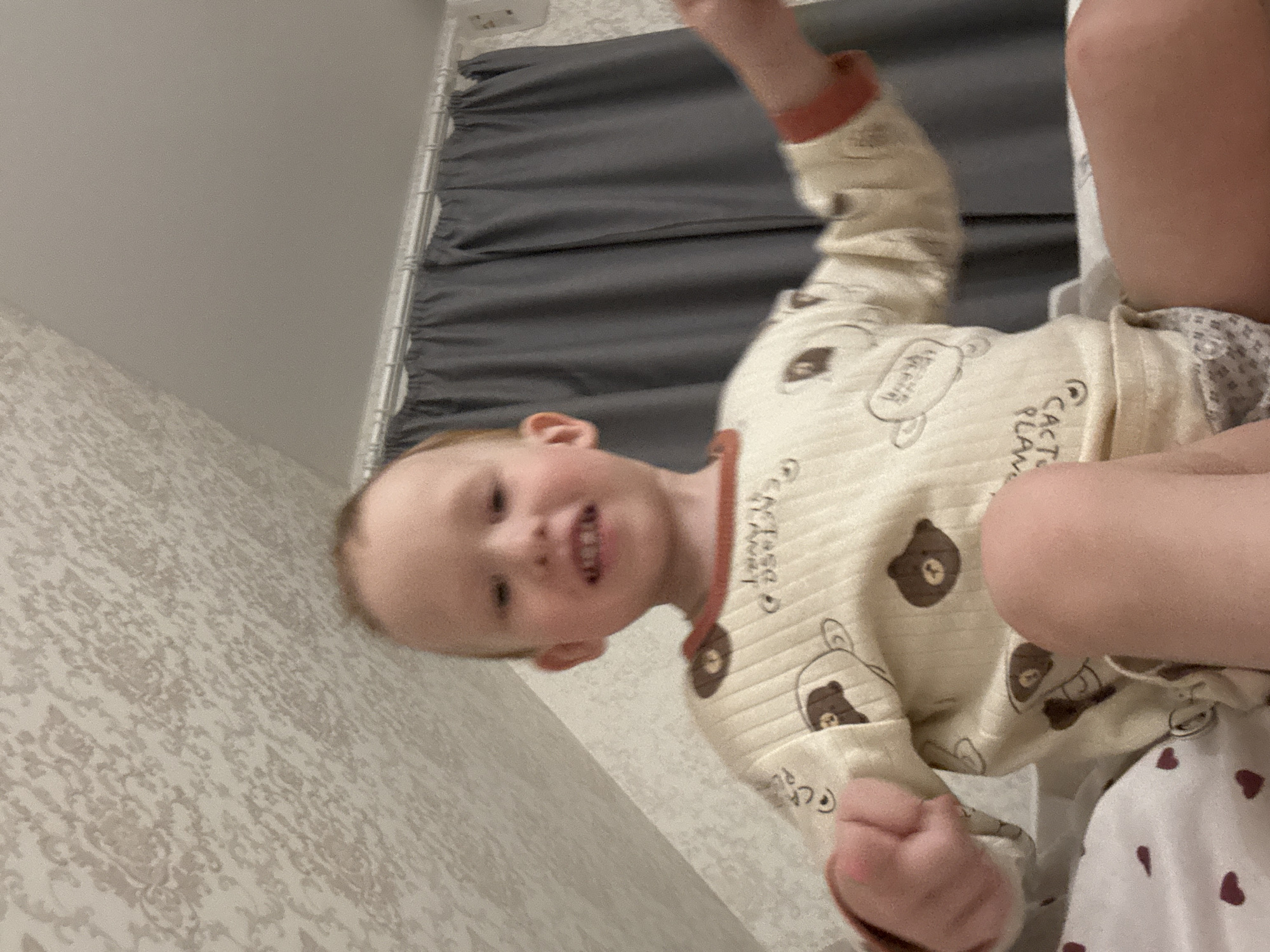 A child, with fair skin and light hair, wearing a beige pajama set with bear and “Cactus Plant” prints, is positioned towards the camera. The child's mouth is open slightly, revealing teeth, and they are raising their arms. The photo is taken from an unusual angle, looking up at the child. The background includes a gray curtain, patterned wallpaper, and a glimpse of white ceiling. The location is likely indoors. The city of Ферма, Russia is not visually identifiable in the image.