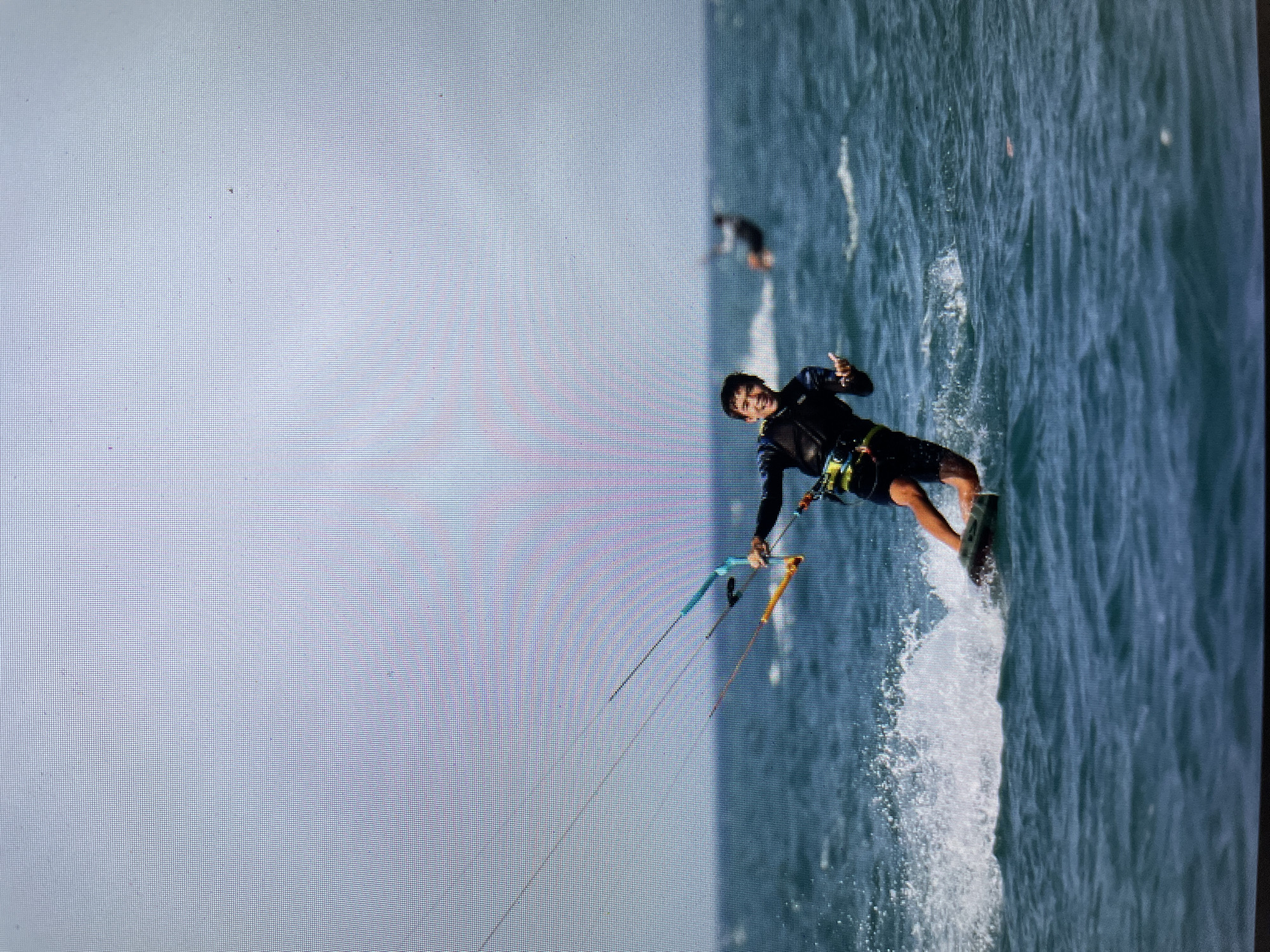 A kitesurfer rides on the water, giving a peace sign with his left hand. He is wearing a dark wetsuit and rides a board. The water around the board is churning with white spray. In the background, another kitesurfer can be seen further out. The sky is overcast. The location is likely in Bacong, Philippines.