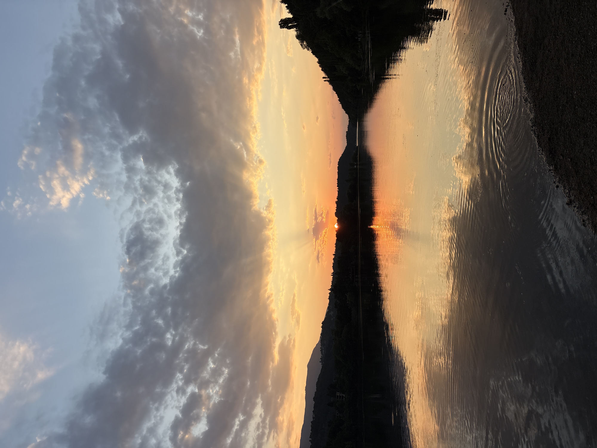 This image depicts a serene sunset over the Pancharevo Lake in Pancharevo, Bulgaria. The sky is a canvas of warm oranges, yellows, and blues, with scattered clouds catching the last rays of the sun. The sun, a bright orange orb, is partially obscured by clouds on the horizon, casting a golden glow across the water. The lake's surface is calm, reflecting the vibrant colors of the sky and the silhouettes of the surrounding trees and hills. The foreground shows a dark, pebbly shore, with gentle ripples disturbing the otherwise still water. The overall scene evokes a sense of tranquility and natural beauty.