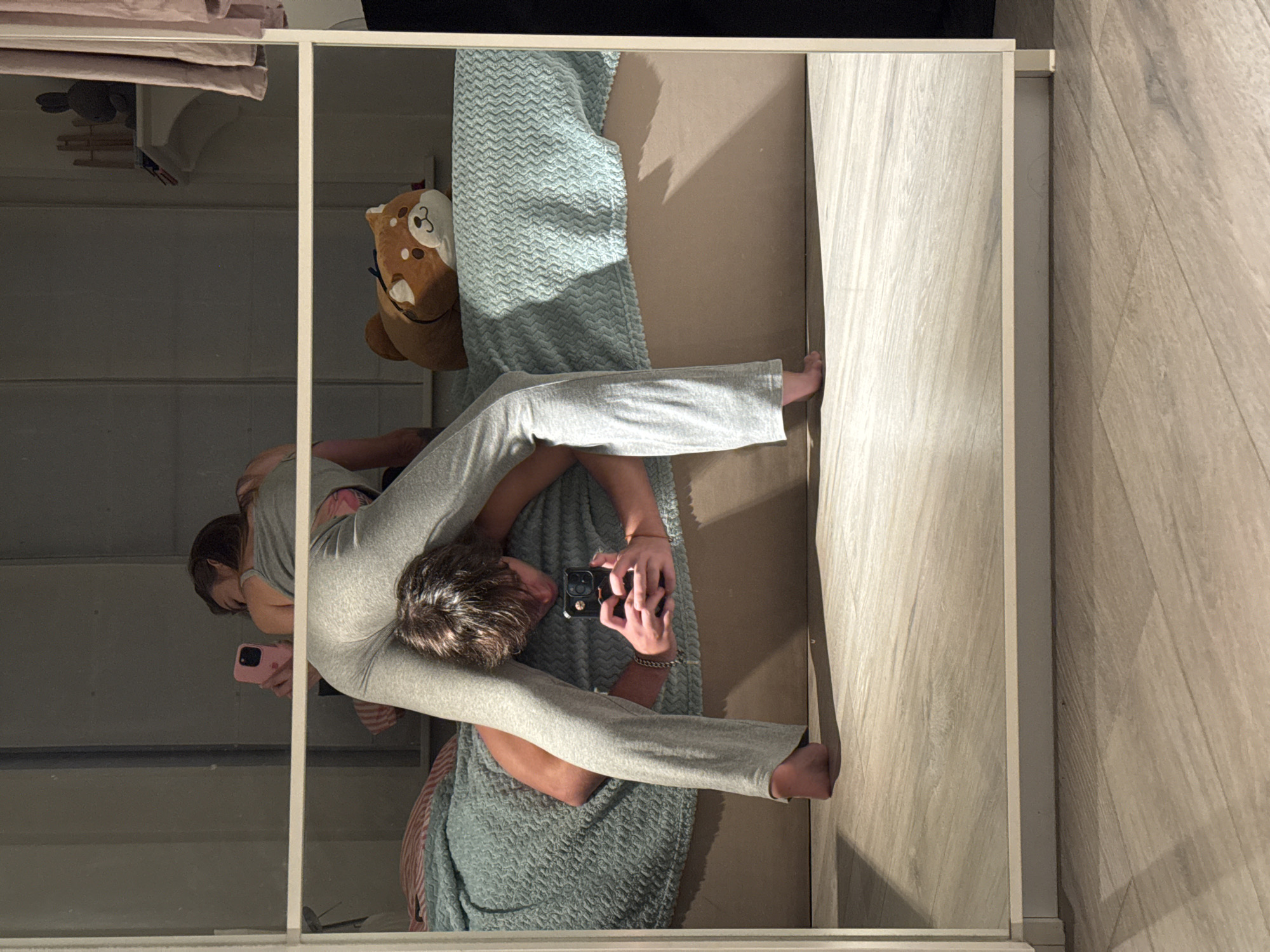 The image depicts a reflection in a mirror of two individuals on a bed. One individual, appearing to be male, is positioned on the bed, facing the mirror and holding a smartphone to take a photograph. This individual is wearing gray sweatpants and is partially naked. The second individual, appearing to be female, is also on the bed, partially visible and holding a pink smartphone. The bed is covered with a light blue textured blanket. A stuffed animal resembling a Shiba Inu dog is visible on the bed. The background of the reflection shows a shelf with a stuffed animal and curtains. The floor in front of the mirror is light-colored wood laminate. The mirror itself is framed in white. The scene appears to be indoors, likely a bedroom. There is no indication of the location being Moscow, Russia.