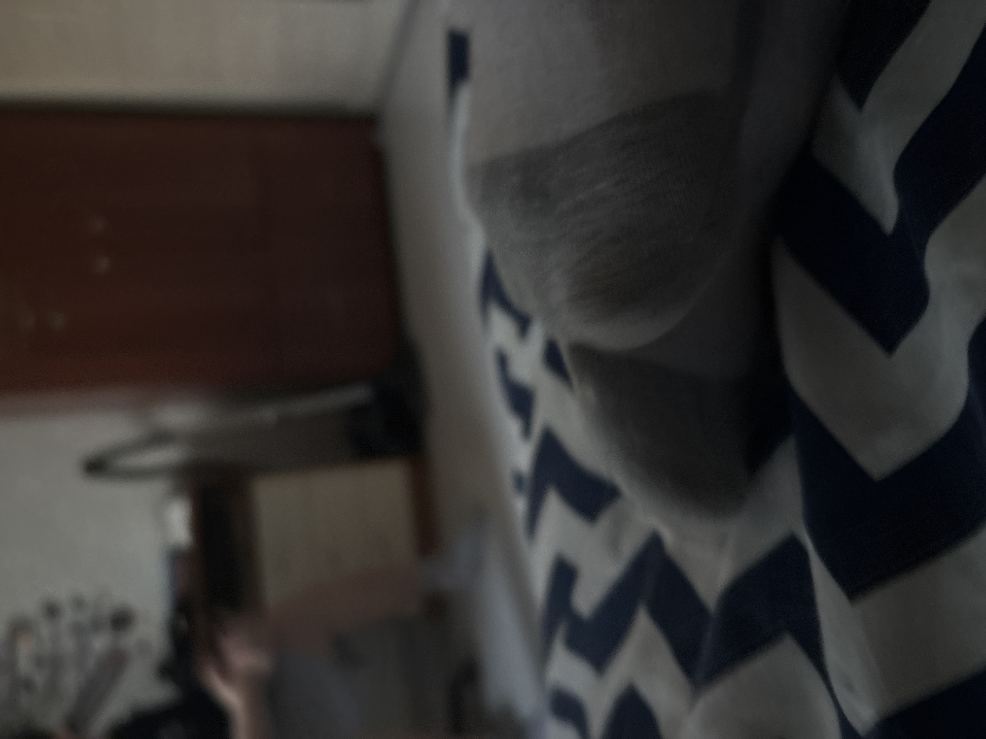 The image is a close-up, blurred, indoor shot. In the foreground, a person's foot in a gray sock is visible, resting on a bed with a blue and white chevron pattern. In the background, a dark reddish-brown wooden dresser or cabinet is present, with a lighter-colored wall to its right. There are also some out-of-focus objects on a shelf or surface to the left, including what appears to be a vacuum cleaner. The lighting is dim.