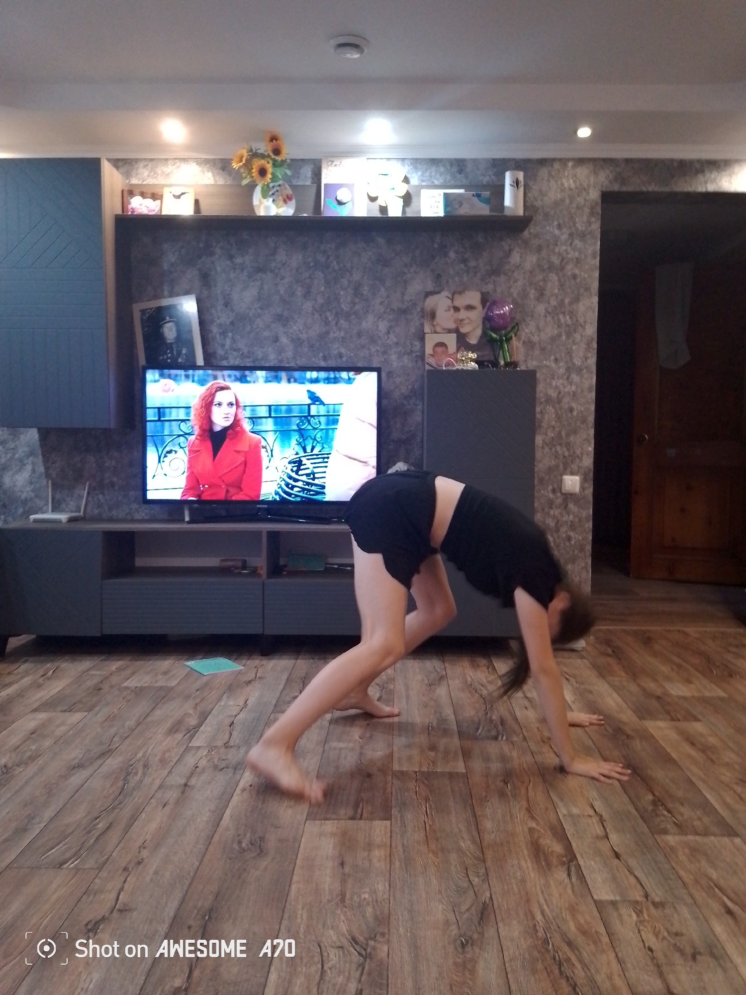 An indoor scene features a young person in black shorts and a black t-shirt performing a yoga pose, with their hands on the wooden floor and their back arched. To the left, a large television displays a woman in a red coat. Above the TV, shelves hold various decorative items, including a vase with sunflowers and framed pictures. The room is furnished with a media console and a cabinet. The floor is made of wood-patterned laminate.