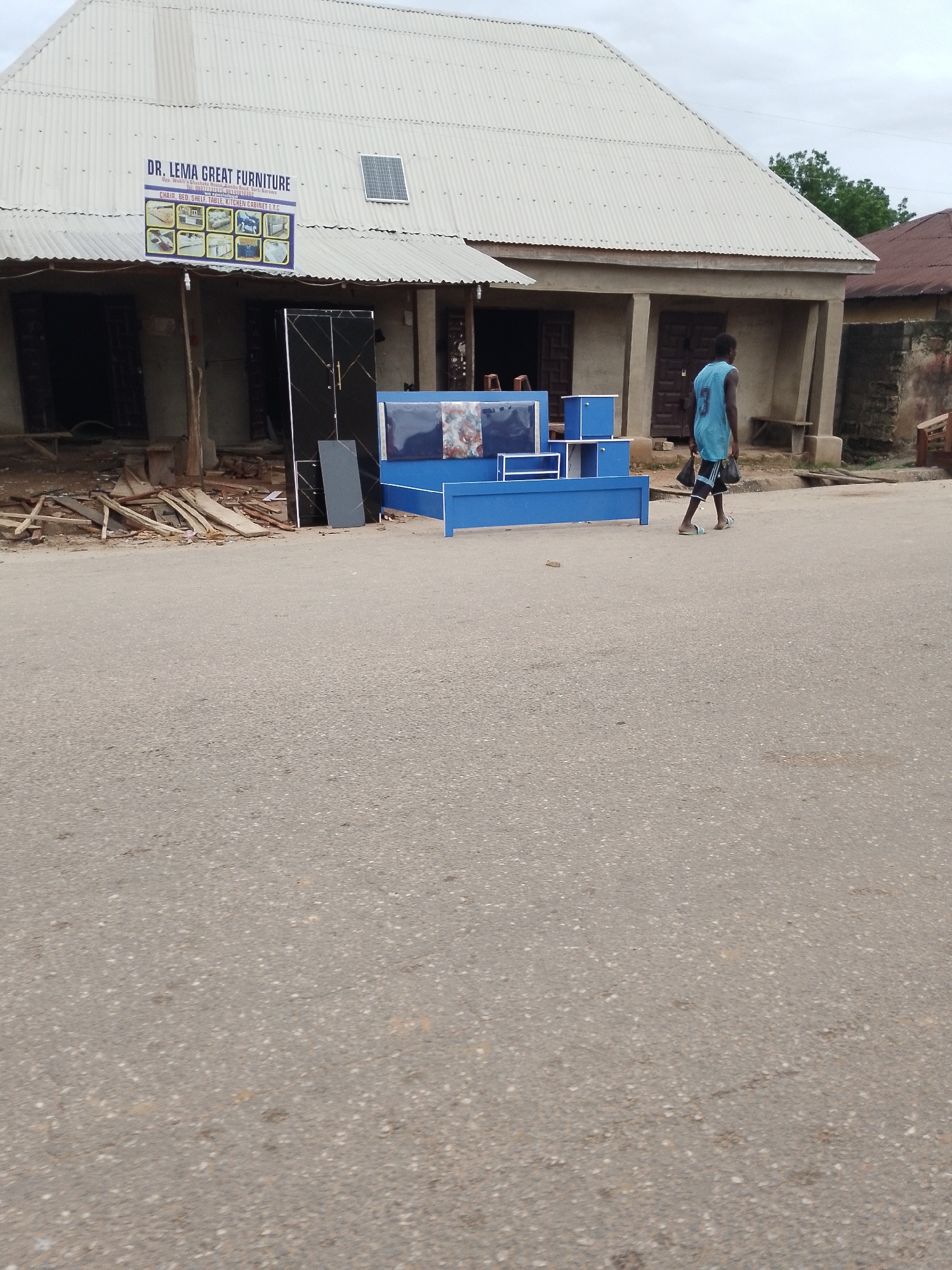 A young man walks on a paved road in front of a corrugated metal building in Serti, Nigeria. The building appears to be a furniture store, with a sign that reads "DR. LEMA GREAT FURNITURE" and shows various furniture items. In front of the building, there is blue furniture, including a bed and storage units. Wood debris is scattered on the ground. The sky is overcast. The man is wearing a light blue tank top and dark shorts, carrying two black bags in his left hand. He is walking away from the camera.