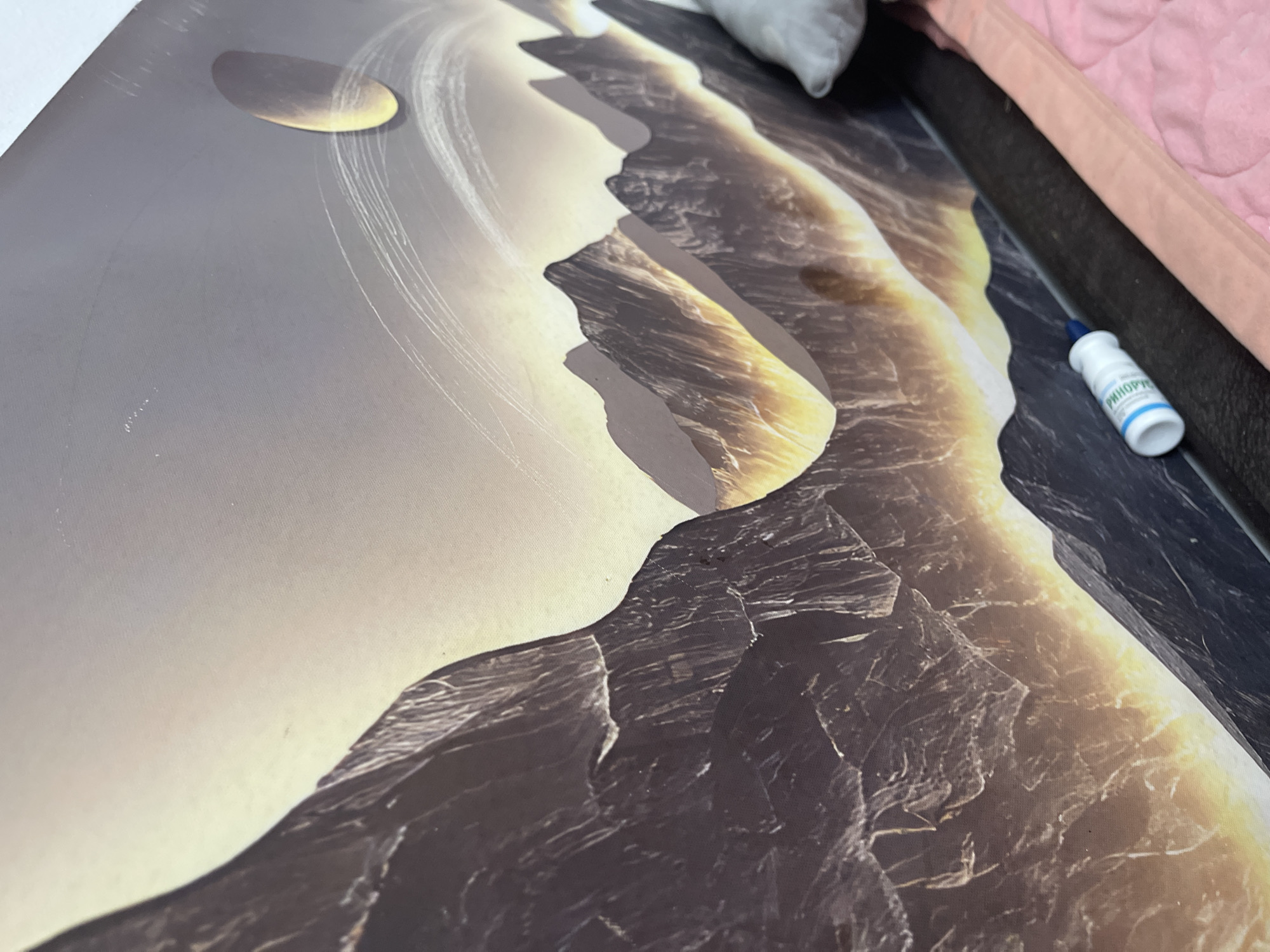A close-up, angled shot of a wall mural depicting a mountainous, possibly alien landscape. The mural is predominantly dark with glowing orange and yellow accents, suggesting a sunset or volcanic activity. A large, oval celestial body, perhaps a planet or moon, is visible in the upper right quadrant of the mural, with faint, arcing lines around it. The mural appears to be printed on a textured surface, possibly canvas or a similar material. In the lower portion of the image, a small white bottle with a blue cap, possibly a nasal spray or eyedropper, lies on a dark surface that appears to be the edge of furniture. A pink textile, possibly a blanket or bedding, is also visible in the lower right corner. The scene is indoors, with no indication of the specific location within Bishkek City, Kyrgyzstan, beyond its presence there.