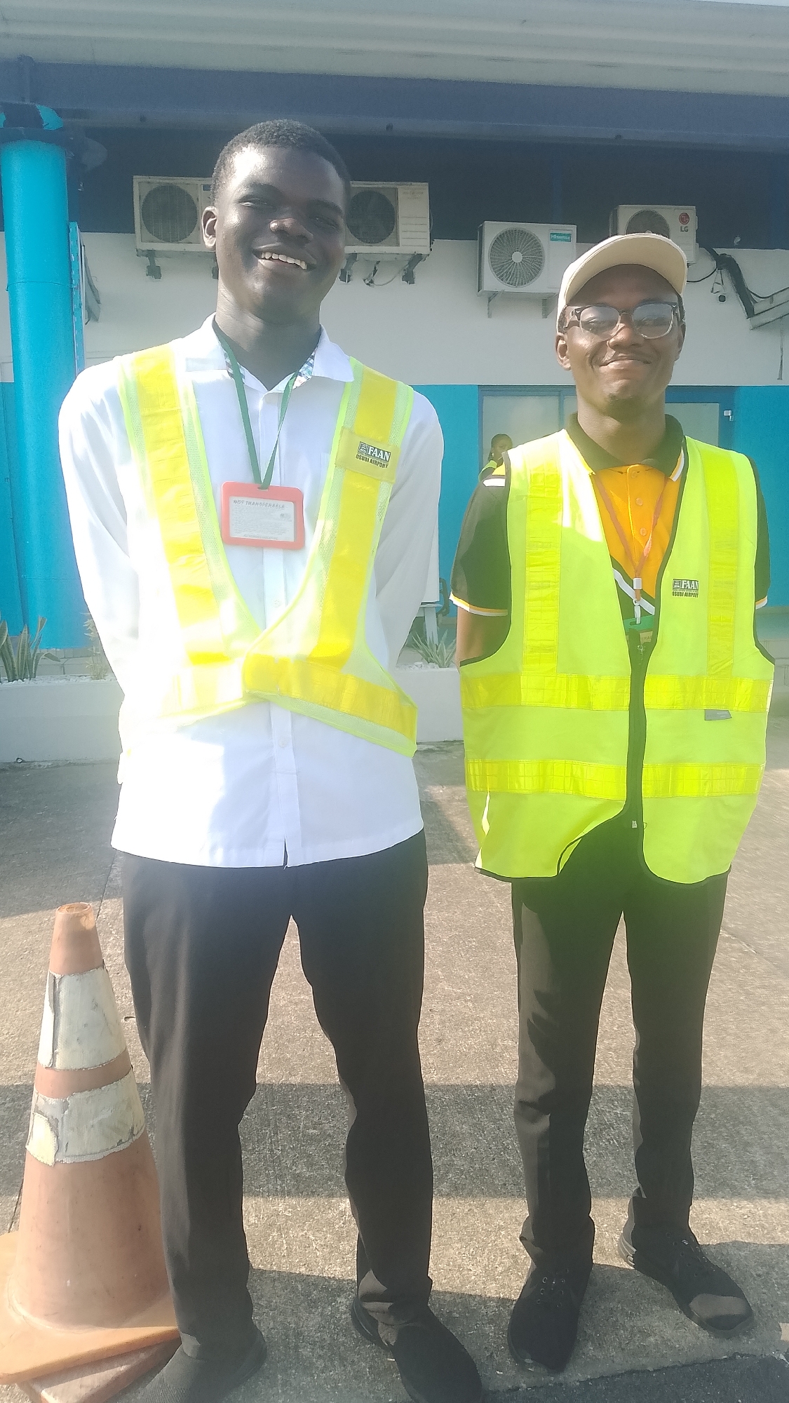 Two young men are standing outside a building in what appears to be an airport environment, likely in Orerokpe, Nigeria, based on the visible "FAAN UGBOKI AIRPORT" logo on their vests. The man on the left is wearing a white collared shirt with a green lanyard and an orange ID card, over which he wears a bright yellow reflective vest. He has a wide, genuine smile. The man on the right is wearing a dark collared shirt with an orange collar, also with a green lanyard and a yellow reflective vest, and a light-colored baseball cap and glasses. He also appears to be smiling. They are standing on a paved surface, with a construction cone to the left of the frame and an air conditioning unit visible on the building behind them. The lighting suggests it is daytime with bright sunlight casting shadows.