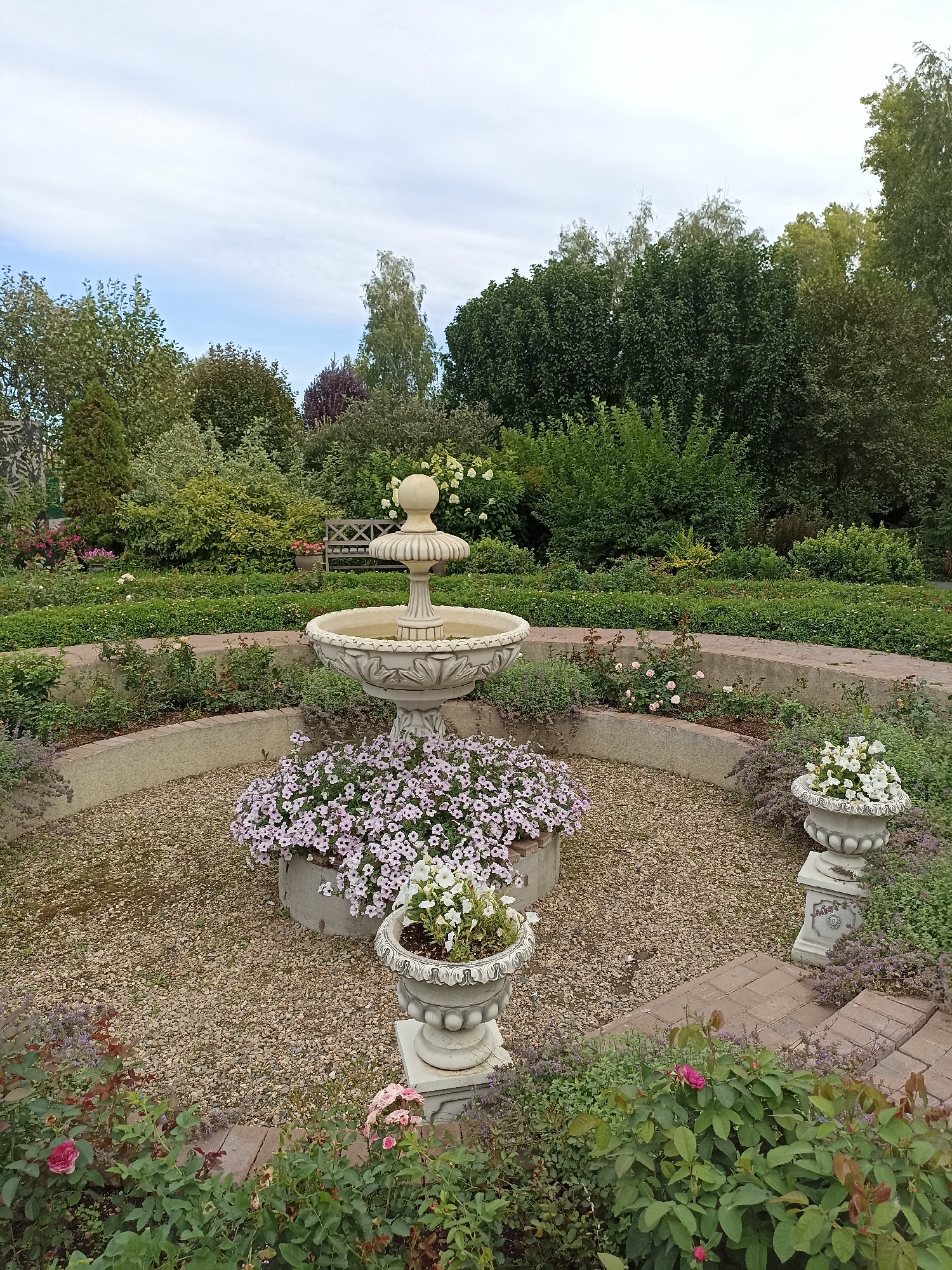 A garden scene with a tiered stone fountain at its center. The fountain is surrounded by a circular gravel path, with planters overflowing with pink petunias and white flowers bordering it. Two ornate stone urns, one containing white flowers and the other an assortment of green plants, are positioned on either side of the gravel area. A curved brick pathway leads further into the garden, where rose bushes with pink and red blooms are visible. In the background, a wooden bench sits amidst lush green trees and shrubs. The image is taken from a slightly elevated perspective, capturing the tranquility of a well-maintained park. The location is the Central Park of Culture and Recreation in Novosibirsk, Russia. There are no people or animals present in the image.