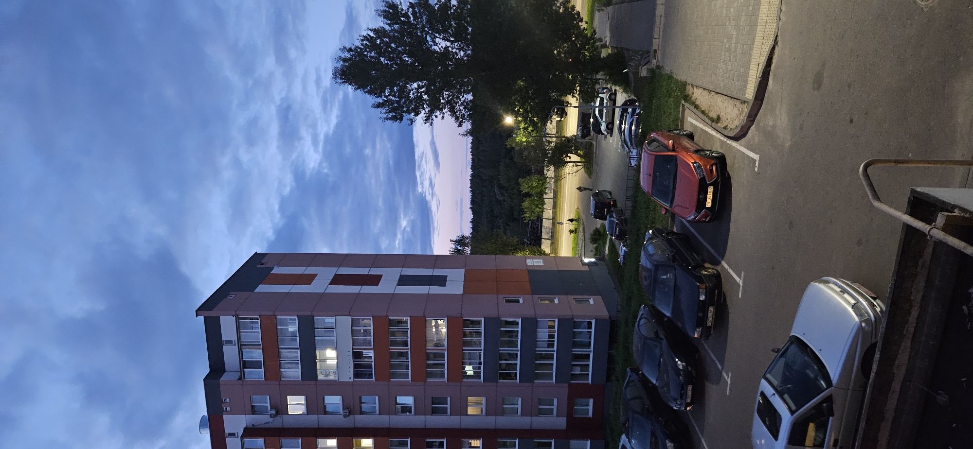 An outdoor, high-angle, vertical, medium shot captures a parking lot in front of a multi-story apartment building under a cloudy twilight sky. The building's facade features a mosaic of rectangular panels in shades of pink, grey, white, and orange. Several cars are parked in the lot. In the foreground, a silver sedan with a sunroof is parked. To its right, a row of parked cars includes a red compact SUV, a dark sedan, and another dark sedan. Further back, a black van and a silver SUV are visible. A large tree stands to the right of the frame, casting shadows. The background reveals a dense line of trees with a faint pinkish hue in the sky beyond them. Several streetlights are illuminated, and two individuals are walking in the distance. The image is situated in the city of Vitsebsk, Belarus.