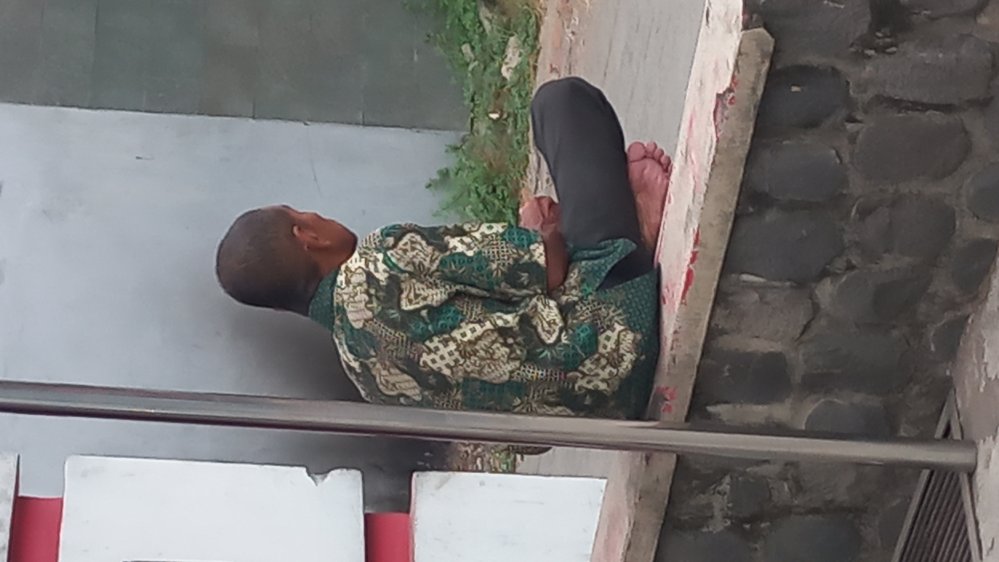 A person sits cross-legged on a stone wall in Pare, Indonesia, facing away from the camera. The individual is wearing a green and white patterned shirt and dark pants. Their bare feet are visible. To the left of the person is a white and red structure, possibly part of a building or sign. A metal pole runs vertically in the foreground, obscuring some of the view. The background consists of a grey wall and some greenery.