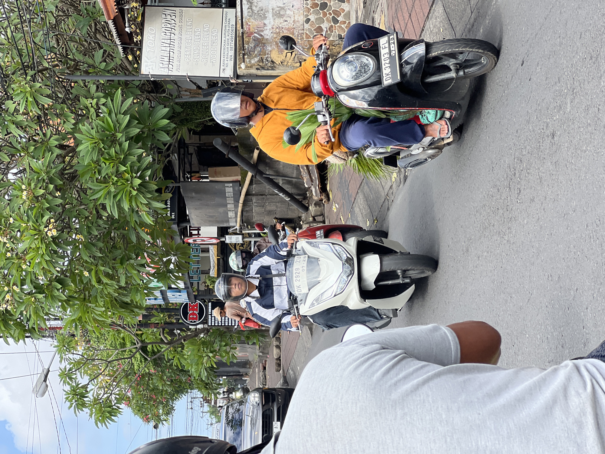 In a street scene in North Kuta, Indonesia, two individuals are riding scooters. The scooter in the foreground, ridden by a person wearing a yellow hooded jacket and blue pants, is black and has a license plate reading "DK 3203 FL." The rider is carrying a bundle of green foliage. Behind this scooter, another person on a white scooter, with a license plate "DK 2928 ZU," is wearing a blue and white striped jacket and dark pants. A grey car is visible in the background, along with trees and buildings lining the street. The sky is overcast.