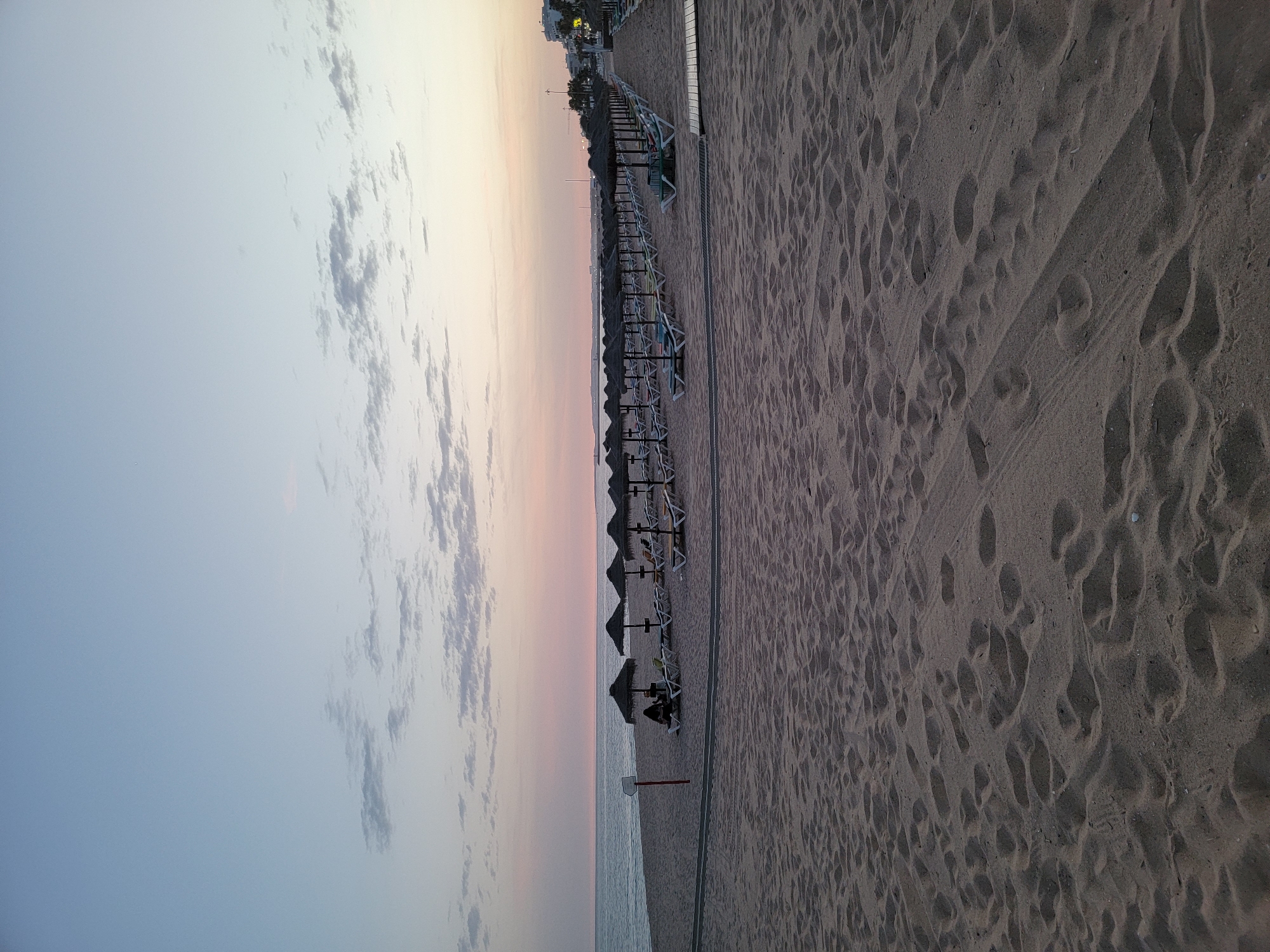 This media file depicts a tranquil beach scene in Quarteira, Portugal, likely during the early morning or late evening. The image is oriented vertically, presenting a panoramic view of the sandy expanse.
The primary subjects include a long row of empty beach chairs and umbrellas lined up along a wooden walkway. The sand is marked with numerous footprints, suggesting recent activity or a busy day prior. In the distance, a few silhouettes of people can be seen relaxing under umbrellas closer to the water's edge. A small, red-topped signpost stands on the sand. The background shows a calm sea meeting the horizon, where the sky transitions from a pale blue to soft hues of pink and orange, indicating either sunrise or sunset. A few distant buildings and palm trees suggest a developed coastal area.
The overall mood is peaceful and serene. The gentle colors of the sky and the absence of strong sunlight contribute to a sense of calm. The presence of the empty chairs and the footprints imply a space designed for leisure and relaxation, now in a quiet phase of the day. The weather appears to be clear and calm, typical of a pleasant coastal environment. No visible text is present in the image.