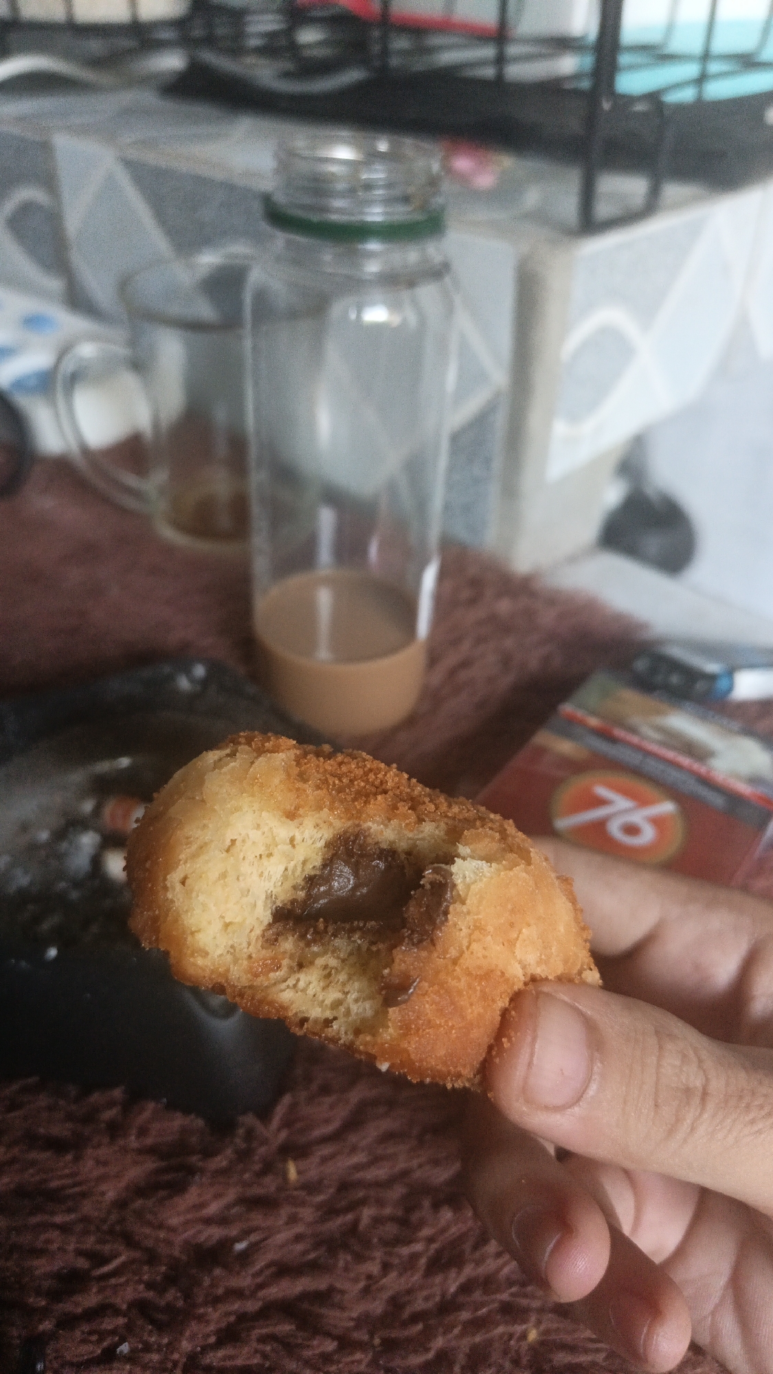 In Tigaraksa, Indonesia, a person is holding a half-eaten pastry filled with chocolate spread. The pastry is golden brown and has a crumbly texture. In the background, a glass bottle with a green cap contains a brown liquid, likely coffee or tea. A glass mug is also visible, along with an ashtray containing cigarette butts. A pack of cigarettes with the "76" logo is partially visible on the right side of the image. The scene appears to be set indoors, possibly on a table covered with a dark, textured cloth.