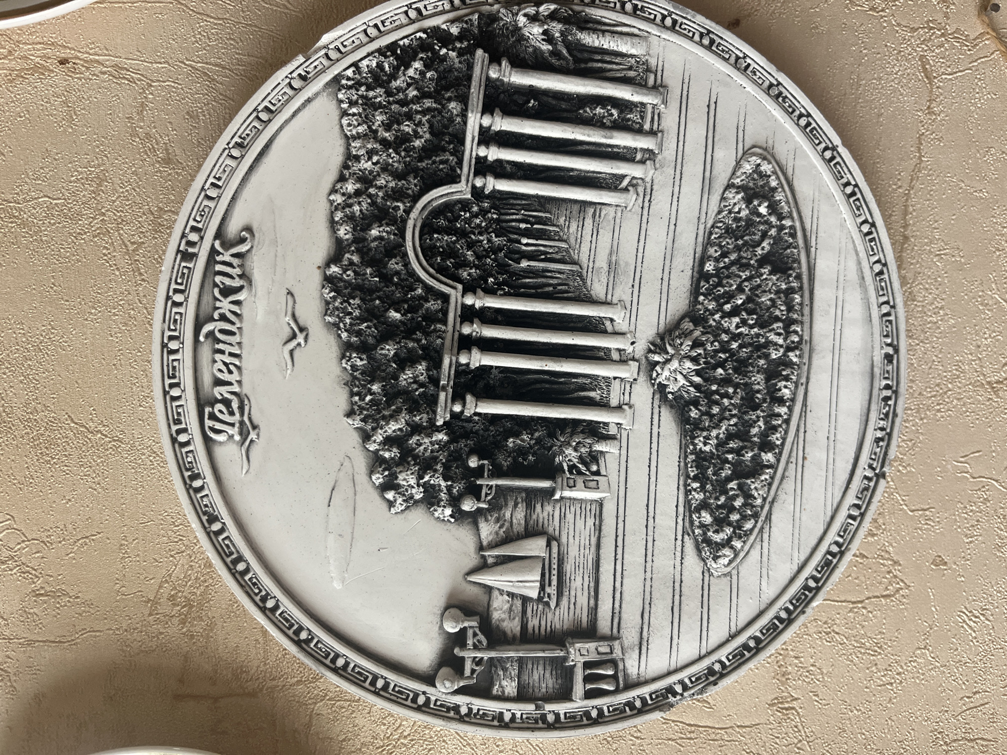 The image shows a circular relief plaque depicting a seaside scene. The plaque is white and mounted on a textured beige wall. Inscribed in Cyrillic script at the top of the plaque is "Геленджик," which translates to Gelendzhik, a resort city on the Black Sea coast of Russia. The scene on the plaque features a body of water with a sailboat, a promenade with lampposts and railings, and classical-style columns forming an archway leading to a treelined path. In the foreground, there is a raised, textured circular island-like feature. The overall style is reminiscent of a souvenir or decorative item.