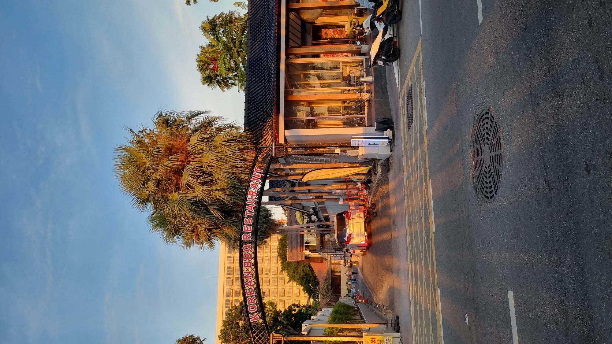 The image shows an outdoor scene, likely a street in Pattaya City, Thailand, captured during sunset, as indicated by the warm, golden light casting long shadows. The sky is a clear, pale blue with a few wispy clouds. Dominating the left side of the frame is a large palm tree with its fronds illuminated by the setting sun.
In the foreground, an archway with decorative metalwork spans across what appears to be a street or pedestrian walkway. Red, illuminated lettering on the arch spells out "KOREAN BBQ RESTAURANT". Behind the arch is a building with a dark, tiled roof, large glass windows, and wooden accents, reflecting the warm light. Several motorcycles are parked in front of the building, one white and black, another yellow.
The street itself is paved and marked with yellow lines. A circular manhole cover is visible on the asphalt. A white van with its brake lights on is driving away from the viewer down the street. Further down the road, other vehicles and a few indistinct figures of people can be seen, suggesting a normal street scene. The overall atmosphere is tranquil and bathed in the soft glow of dusk. The warm lighting and the presence of palm trees contribute to a tropical, evening ambiance.