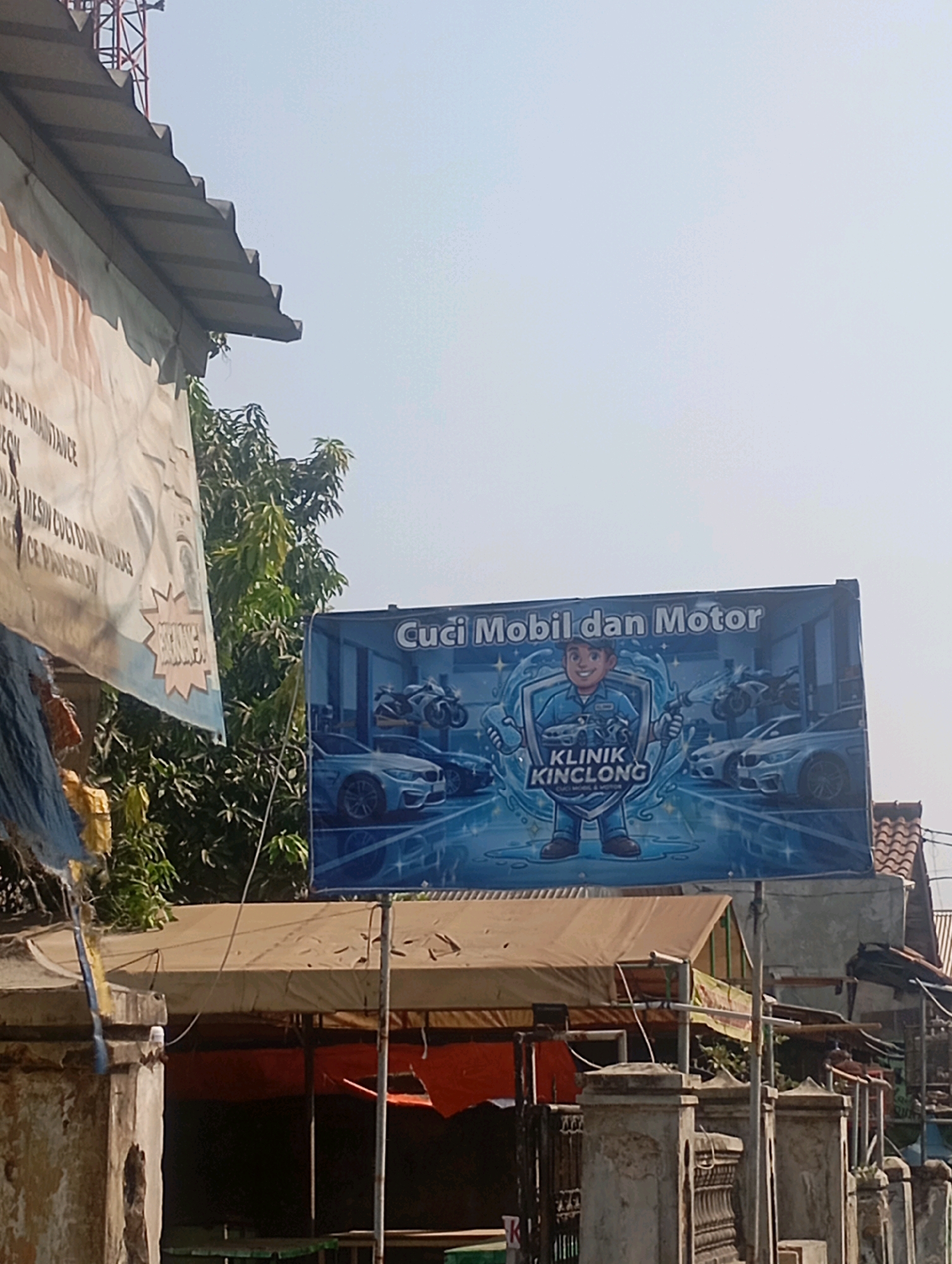 An outdoor scene in Babelan, Indonesia, features a bright blue billboard advertising "Cuci Mobil dan Motor" (Car and Motorcycle Wash) by "KLINIK KINCLONG" (Sparkling Clinic). The billboard depicts a cartoon character holding a hose, surrounded by sparkling water, with images of cars and motorcycles. To the left, a torn white banner with text in Indonesian, possibly listing services, is partially visible, hanging over a corrugated metal roof. Below the billboard, a covered structure with orange tarps and visible tables suggests an outdoor seating or workspace. The background shows trees, buildings with tiled roofs, and a clear sky. The foreground is dominated by concrete pillars and fences.