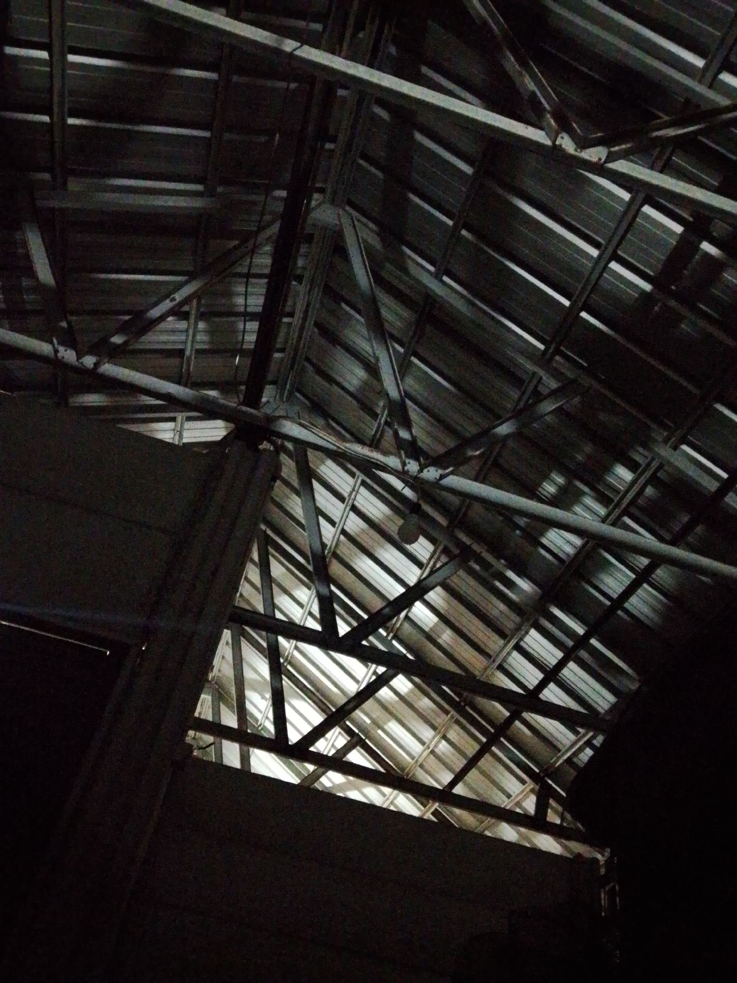 The image shows a dimly lit interior view, likely taken inside a building or structure in Bukit Ketok, Indonesia. The focus is on the ceiling, which is constructed of corrugated metal sheets supported by a network of steel beams and trusses. Gaps in the roofing allow for some light to penetrate, creating areas of brightness against the otherwise dark and shadowed structure. A hanging light bulb is visible amidst the structural elements. The walls of the building are dark and indistinct.