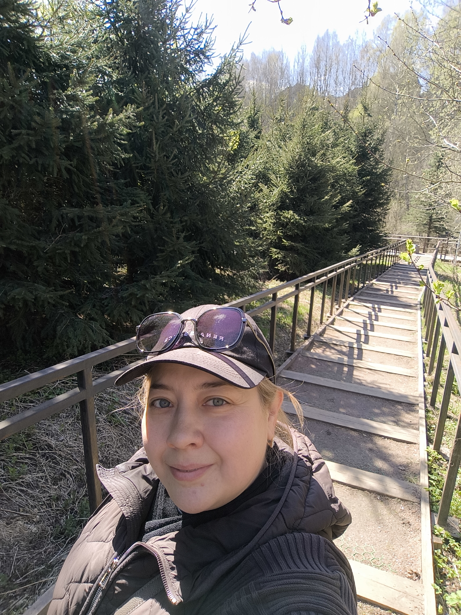 A selfie-style portrait captures a woman outdoors on a bright, sunny day. She is positioned in the foreground, looking directly at the camera with a calm, pleasant expression. She has light-colored hair, blue-grey eyes, and is wearing a dark puffer jacket over a dark ribbed sweater or scarf. A black baseball cap sits on her head, topped with dark sunglasses that reflect the reversed text "BENICE" and other indistinct characters.
Behind her, a long, wide wooden staircase with sturdy metal railings on both sides descends down a slope. Strong shadows from the railings are cast across the steps, indicating bright sunlight, likely mid-morning or early afternoon. To the left of the staircase, dense evergreen trees (spruces or firs) create a lush backdrop. Further in the background, deciduous trees appear to be in early spring, with many branches still bare or just beginning to bud, under a clear blue sky. The scene suggests a scenic park or natural pathway in Moscow, Russia.