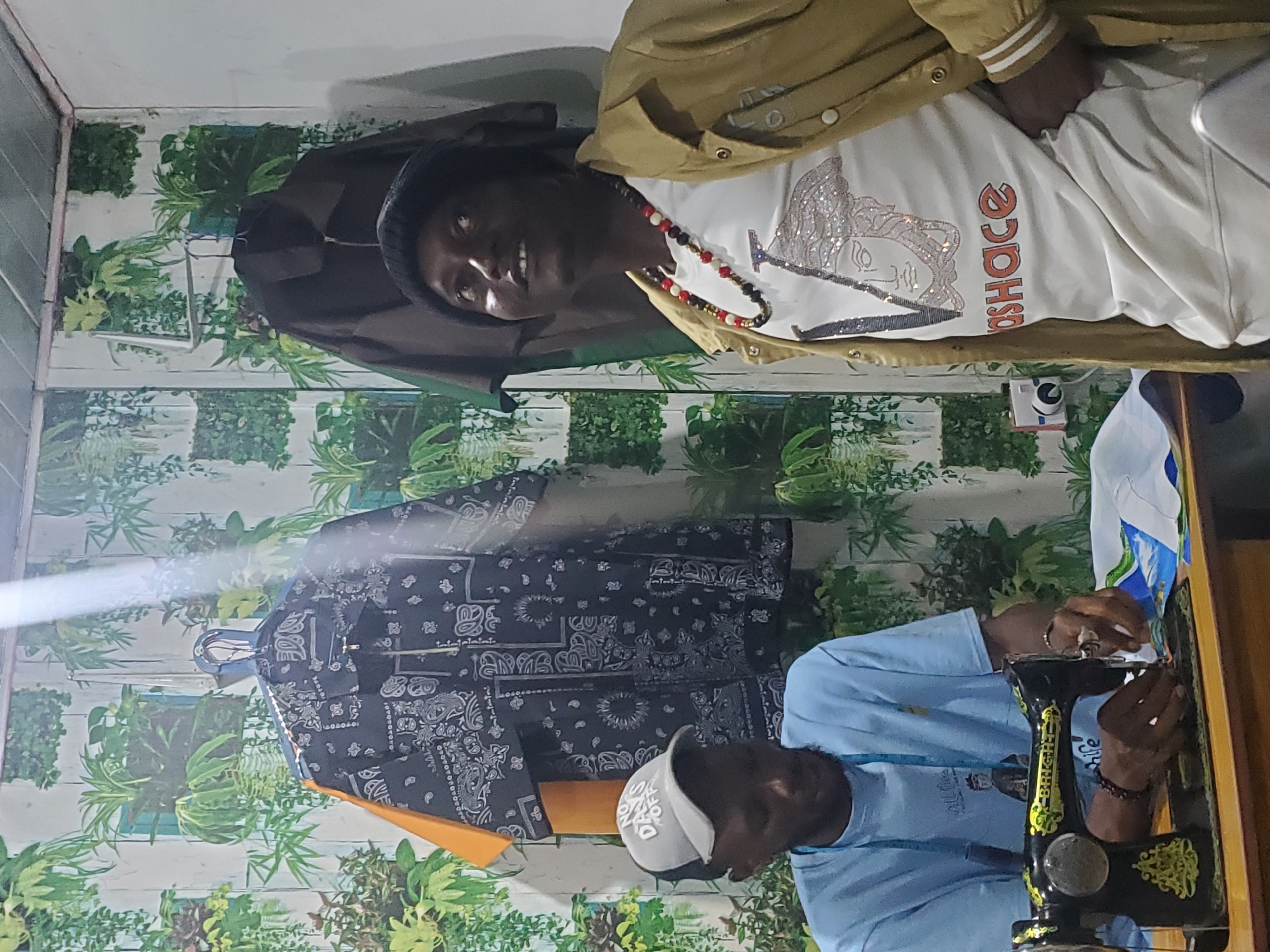 The image shows two individuals in what appears to be a tailoring workshop in Jos, Nigeria. The scene is vibrant, with a backdrop of artificial plants and a white wooden-paneled wall, suggesting an indoor setting.
The subject in the foreground, wearing a light blue long-sleeved shirt and a white baseball cap, is actively engaged in sewing. They are seated at a black sewing machine with yellow decorative accents. Their hands are positioned over the fabric and the needle, indicating concentration on their task. A blue measuring tape is draped around their neck, a common tool for tailors.
Behind the sewer, another individual is visible, wearing a tan jacket over a white t-shirt. The t-shirt has a striking design featuring a portrait embellished with what looks like glitter or rhinestones, and text that reads "VERSACE." They are also wearing a beaded necklace. This person is looking towards the camera and appears to be posing.
On a hanger in the background, a black garment with a white paisley pattern is displayed. This garment, along with the presence of the sewing machine and the tailor at work, strongly suggests that the activity taking place is garment creation or alteration.
The lighting in the image is a bit diffused, but it doesn't clearly indicate the time of day or weather conditions outside. The overall mood seems to be one of focused work and a creative environment. The presence of artificial plants might be a design choice to bring an outdoor feel indoors, or to enhance the aesthetic of the workshop.