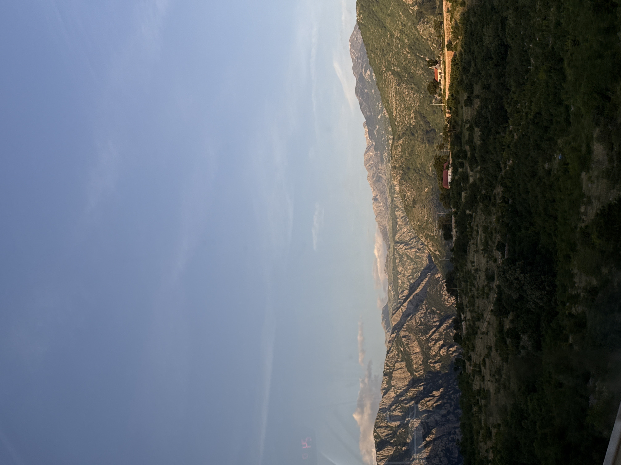 The image captures a view of a mountainous landscape in or near Klopot, Montenegro. The scene is dominated by rugged, rocky mountains covered with sparse vegetation and denser forests closer to the foreground. A few buildings are visible along the mountain slopes. The sky is blue with wisps of clouds. The image appears to be taken from an elevated position, possibly from a cable car or viewing platform. The orientation of the image is sideways.