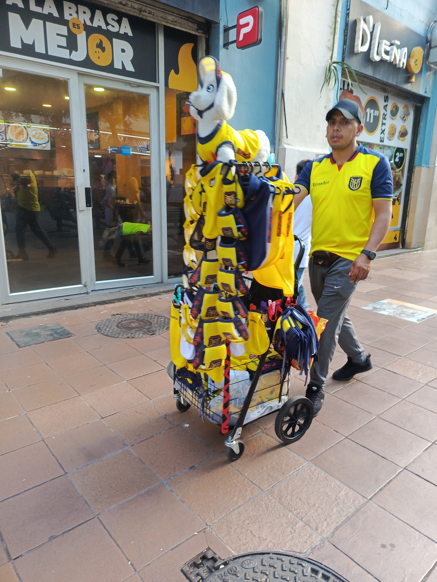 In Guayaquil, Ecuador, a man wearing a yellow and blue Ecuadorian soccer jersey, dark baseball cap, and grey sweatpants walks past a cart laden with yellow merchandise, including replicas of the Ecuadorian national soccer team's jersey and other fan apparel. The cart, a multi-tiered structure with wheels, is positioned on a tiled sidewalk in front of a storefront. The storefront features large glass windows displaying food items and has signage that reads "A LA BRASA ES MEJOR." In the background, other individuals can be seen through the glass windows. The overall scene depicts a commercial street environment.