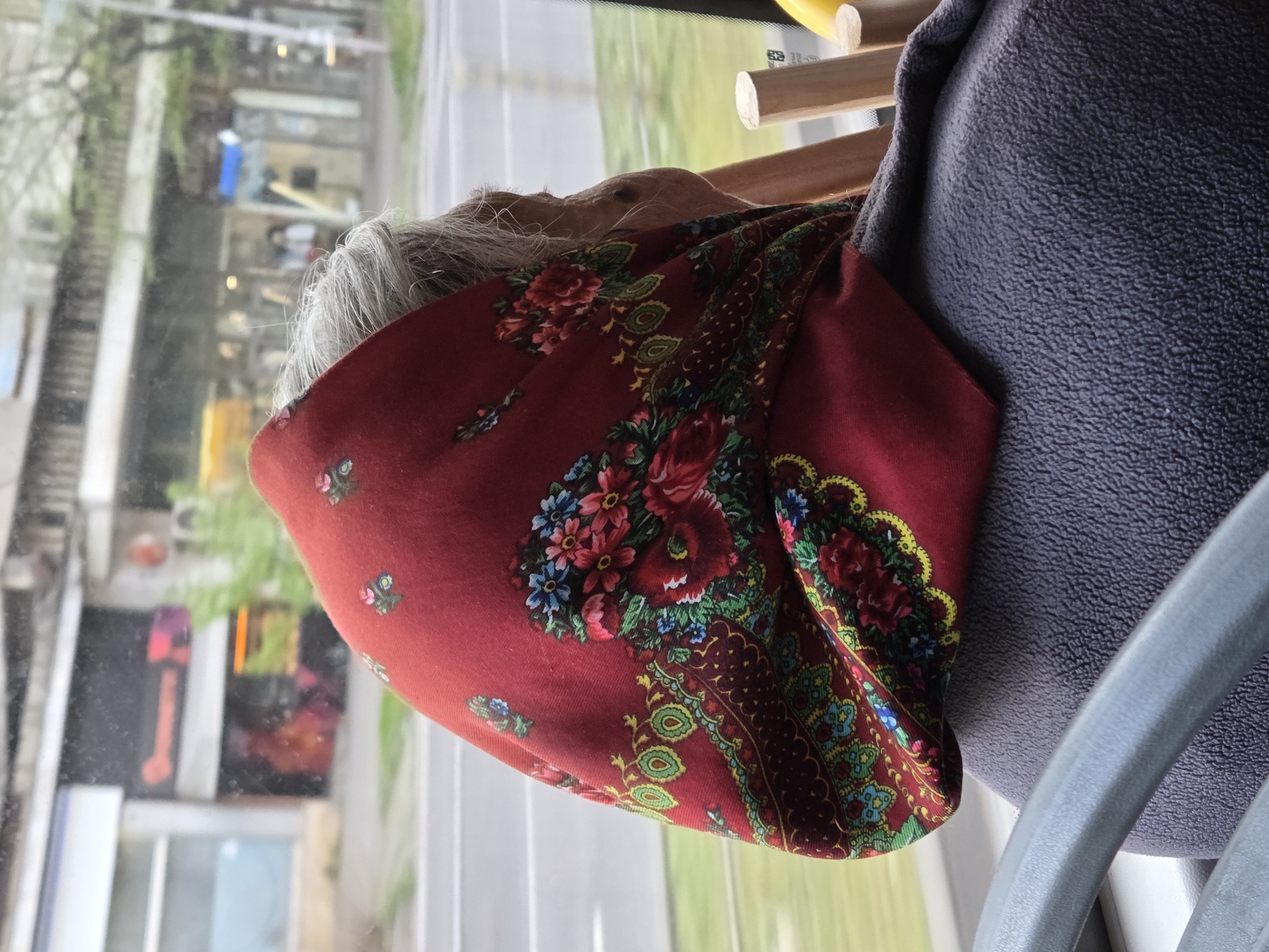 An elderly person, likely a woman, is seated inside a moving vehicle, possibly a bus or tram, facing away from the viewer and looking out a window. Her grey hair is visible around her head, covered by a vibrant, traditional red headscarf, often referred to as a babushka. The headscarf features intricate floral patterns in shades of red, pink, blue, green, and yellow, with dark borders and motifs.
To her right, a grey or dark blue textured fabric, likely a seat or backrest, is visible. Above her head, there are wooden slats, possibly part of an overhead luggage rack or interior paneling of the vehicle. A grey plastic or metal handle/bar is seen in the foreground on the right.
Through the large window, a blurred urban street scene passes by, indicating the vehicle is in motion. Buildings, trees, and a road are discernible, along with some indistinct shapes and lights. The outdoor lighting suggests it is daytime, possibly overcast, contributing to a muted, soft illumination. The scene depicts a street in Varna, Bulgaria. No legible text is clearly visible on the headscarf or any prominent objects within the frame, though a small, unreadable sticker appears on the window frame in one of the cropped images. The overall impression is one of quiet travel through a city.