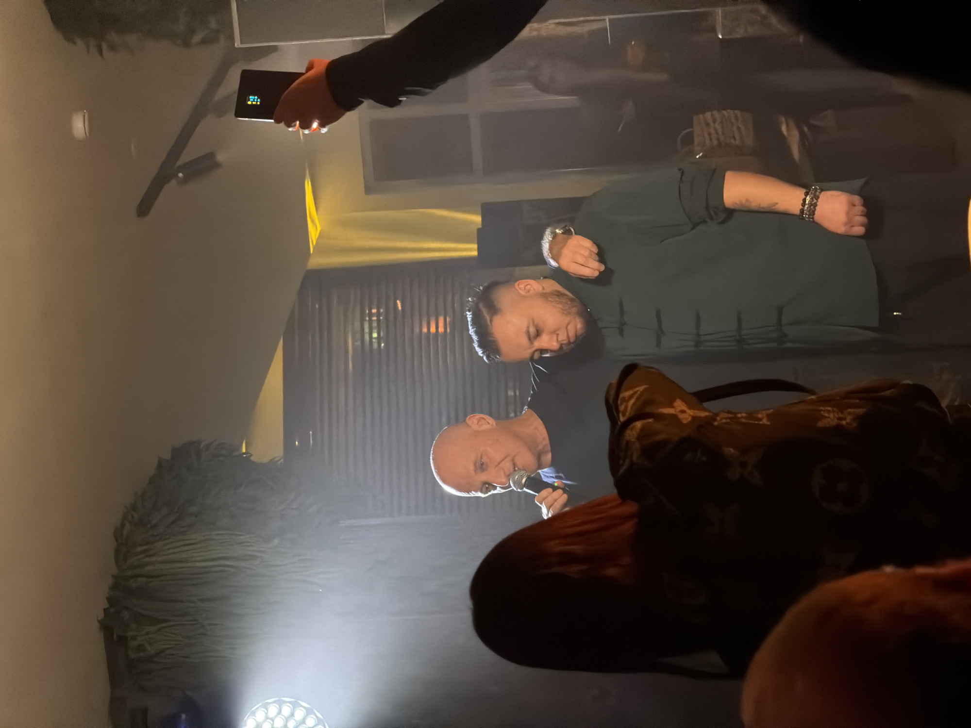 Two men are on a stage. One is bald and holding a microphone. The other is wearing a green shirt. Someone is holding a phone. The location is in Moscow, Russia.